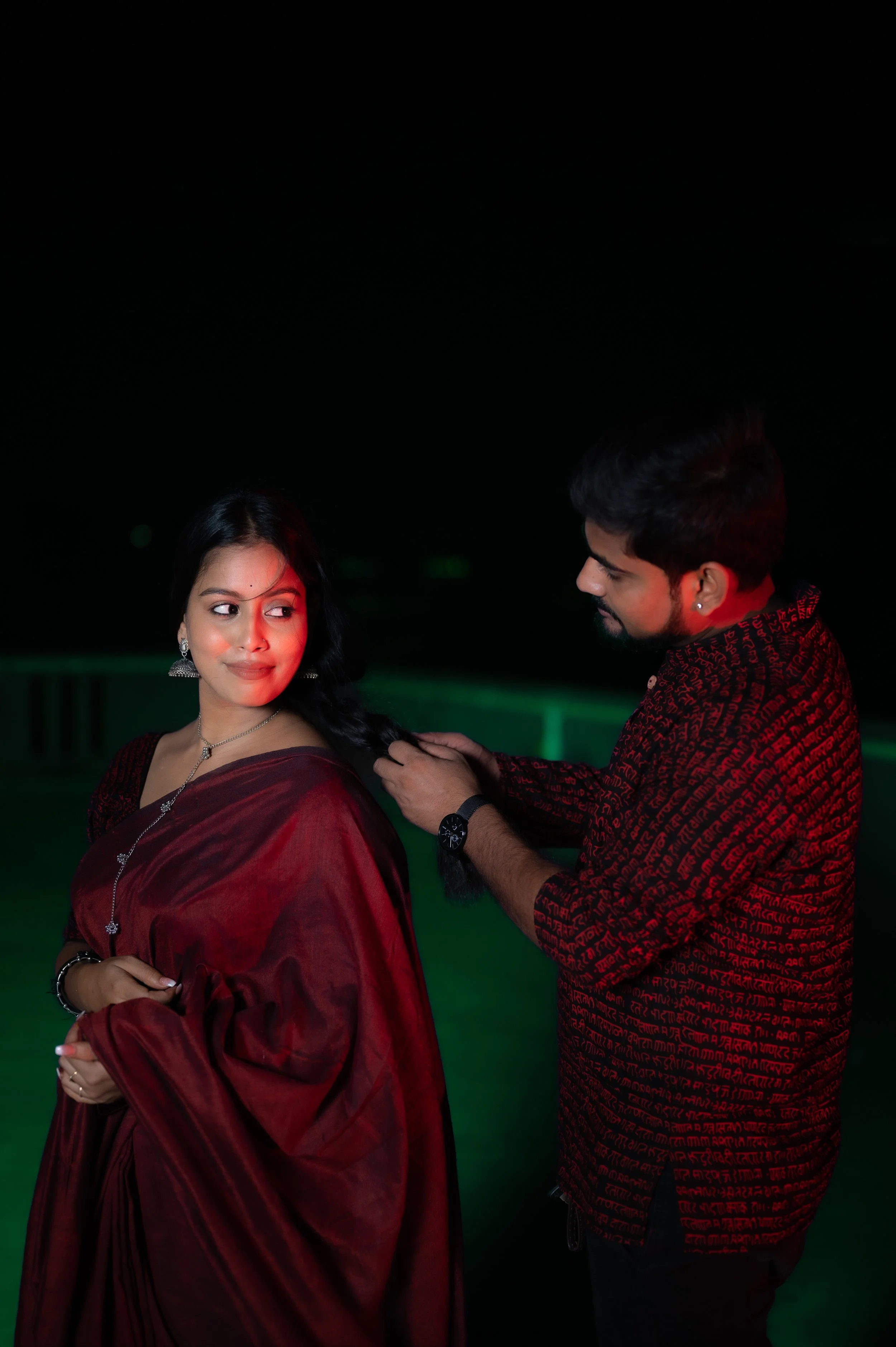 A man helping a woman with her long black hair on a rooftop at night, both wearing dark red and black clothes, illuminated by red and green lighting.
