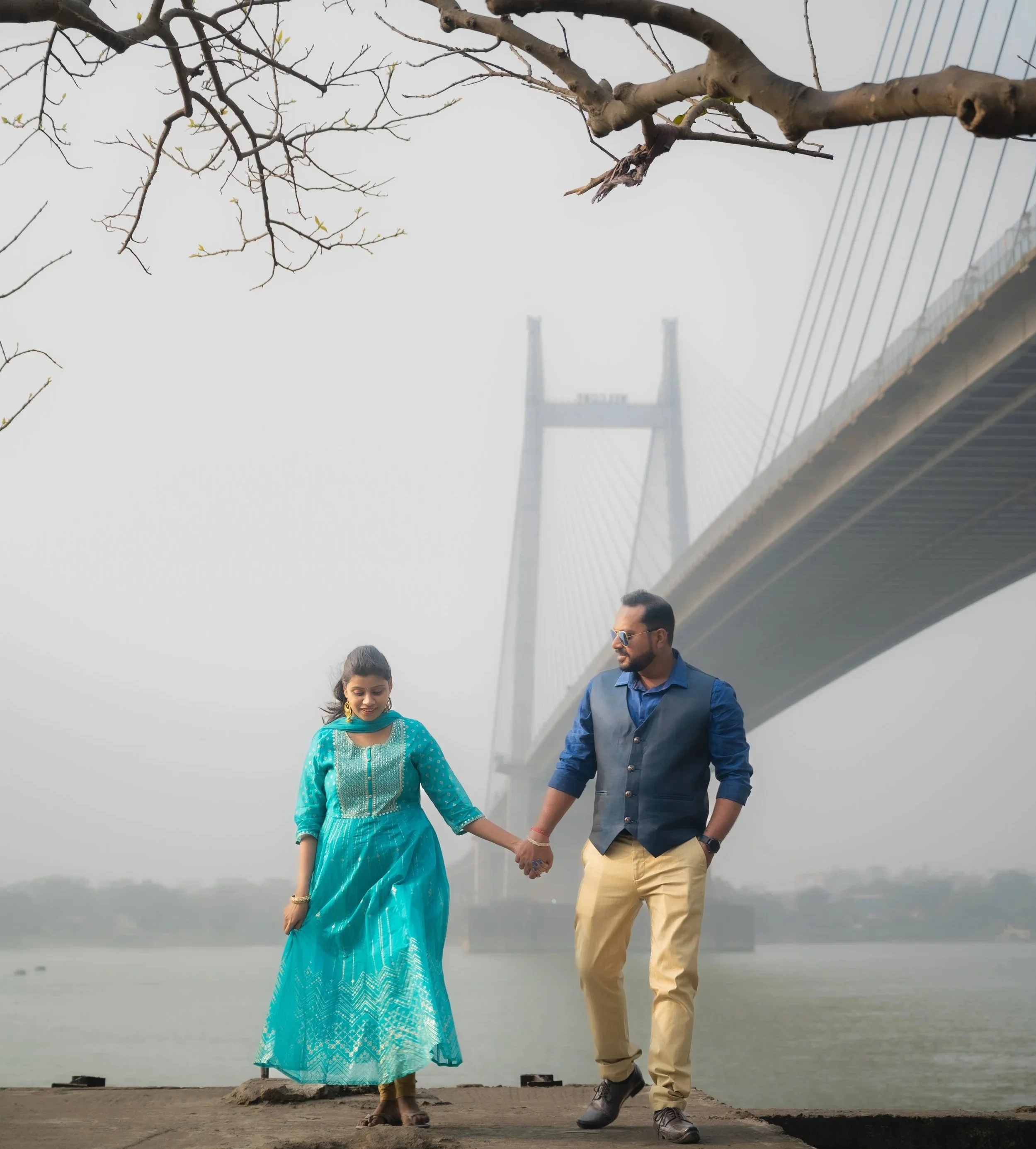 A couple holding hands, walking together on a foggy day near a body of water, with a bridge in the background. The woman is wearing a bright blue traditional dress, and the man is dressed in casual beige pants and a blue shirt.