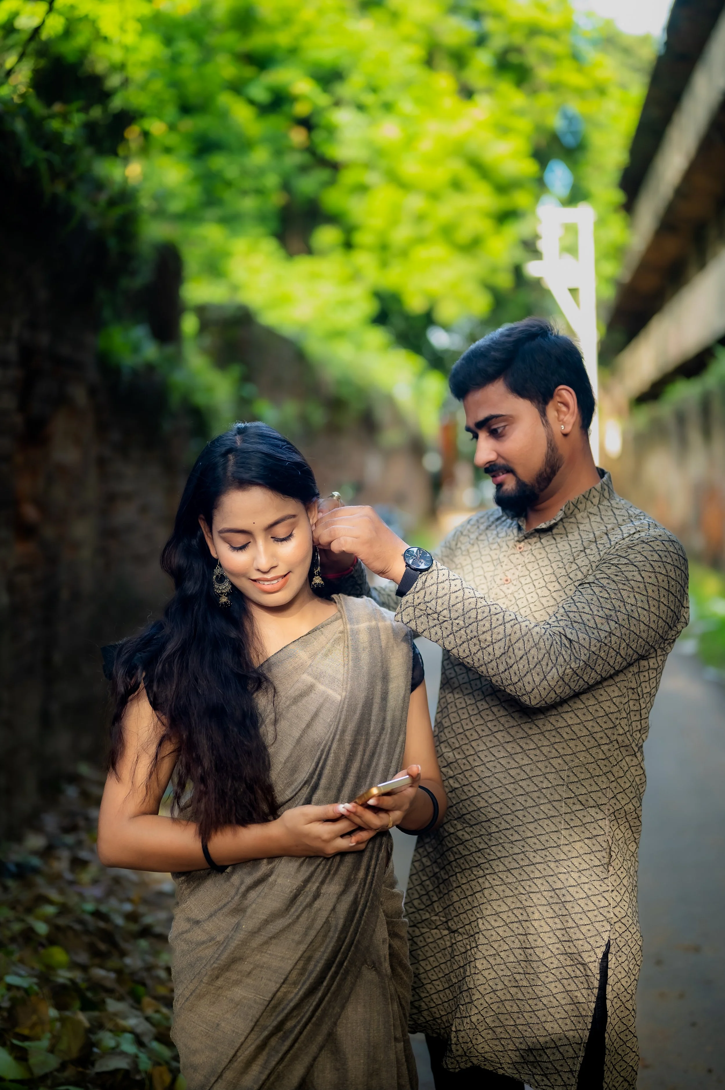 A man helping a woman with her earrings outdoors on a sunny day with green trees in the background.