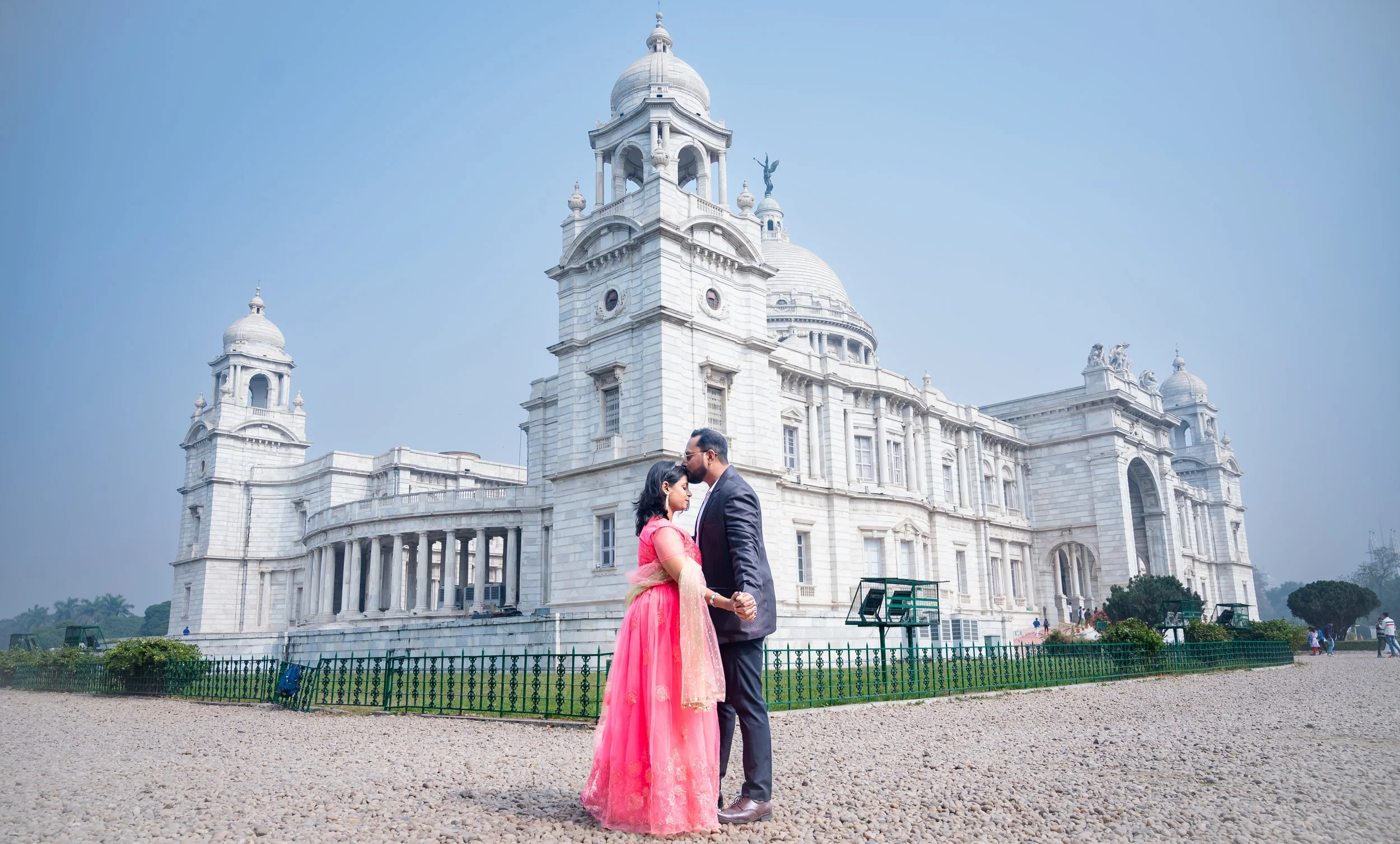 A couple dressed in formal Indian attire stands close together in front of the Victoria Memorial in Kolkata, India, with the man gently kissing the woman's forehead, against a clear sky.