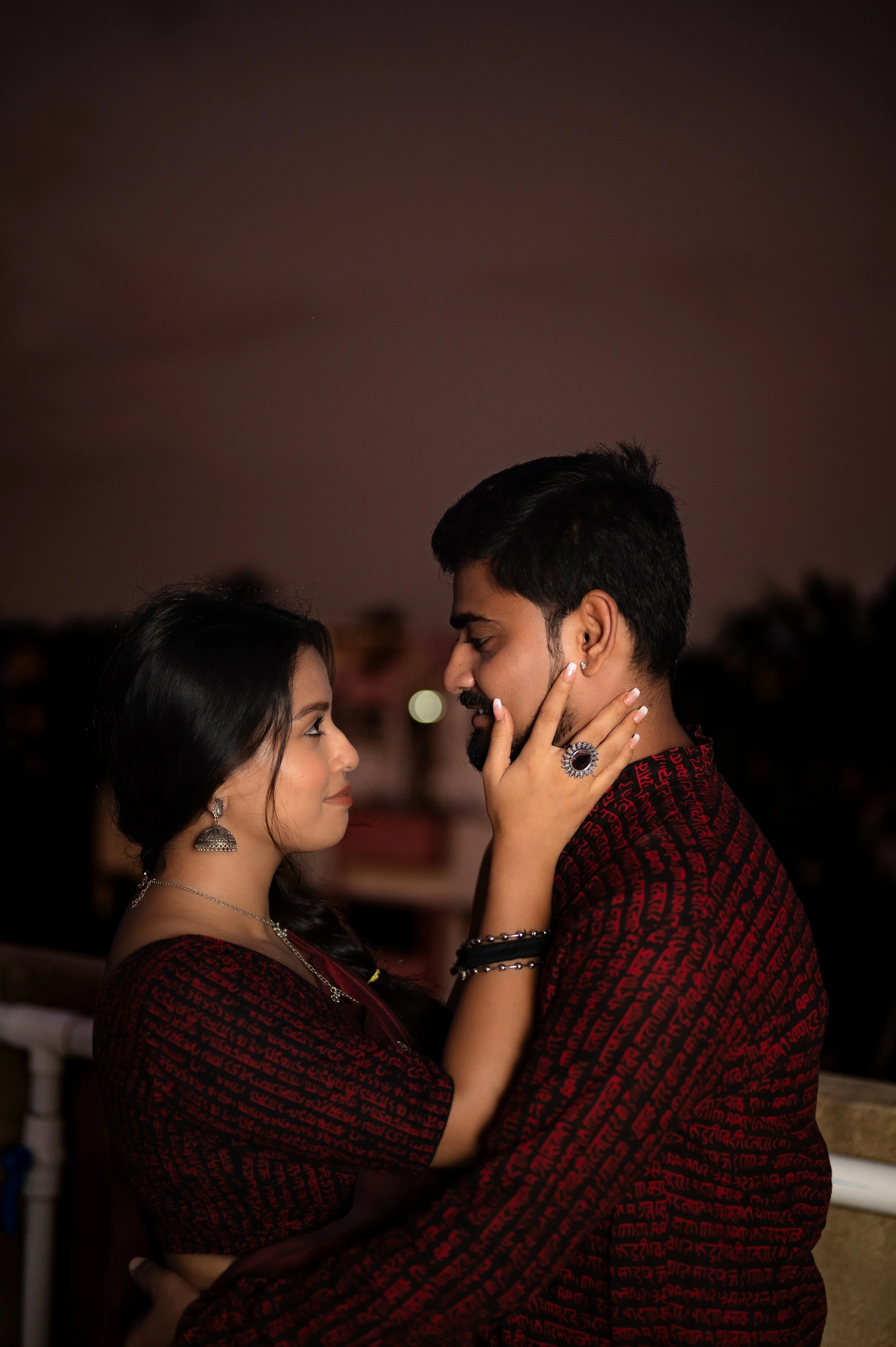 A couple dancing closely at night with a dark sky and blurred city lights in the background, dressed in matching maroon and black patterned outfits, gazing into each other's eyes.