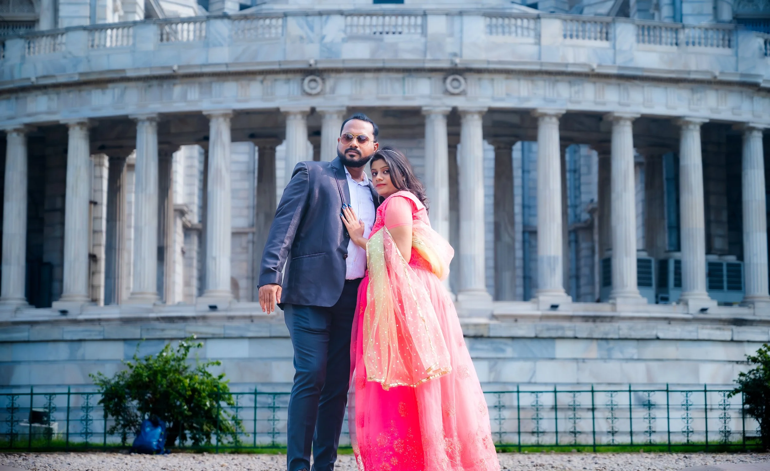 A man wearing sunglasses and a suit jacket standing next to a woman in a pink and gold traditional dress, in front of a large stone building with columns.
