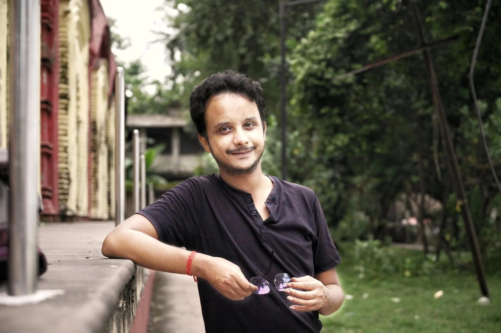 A young man with dark hair, wearing a black shirt, sitting outdoors near a park railing, holding sunglasses, with green trees in the background.