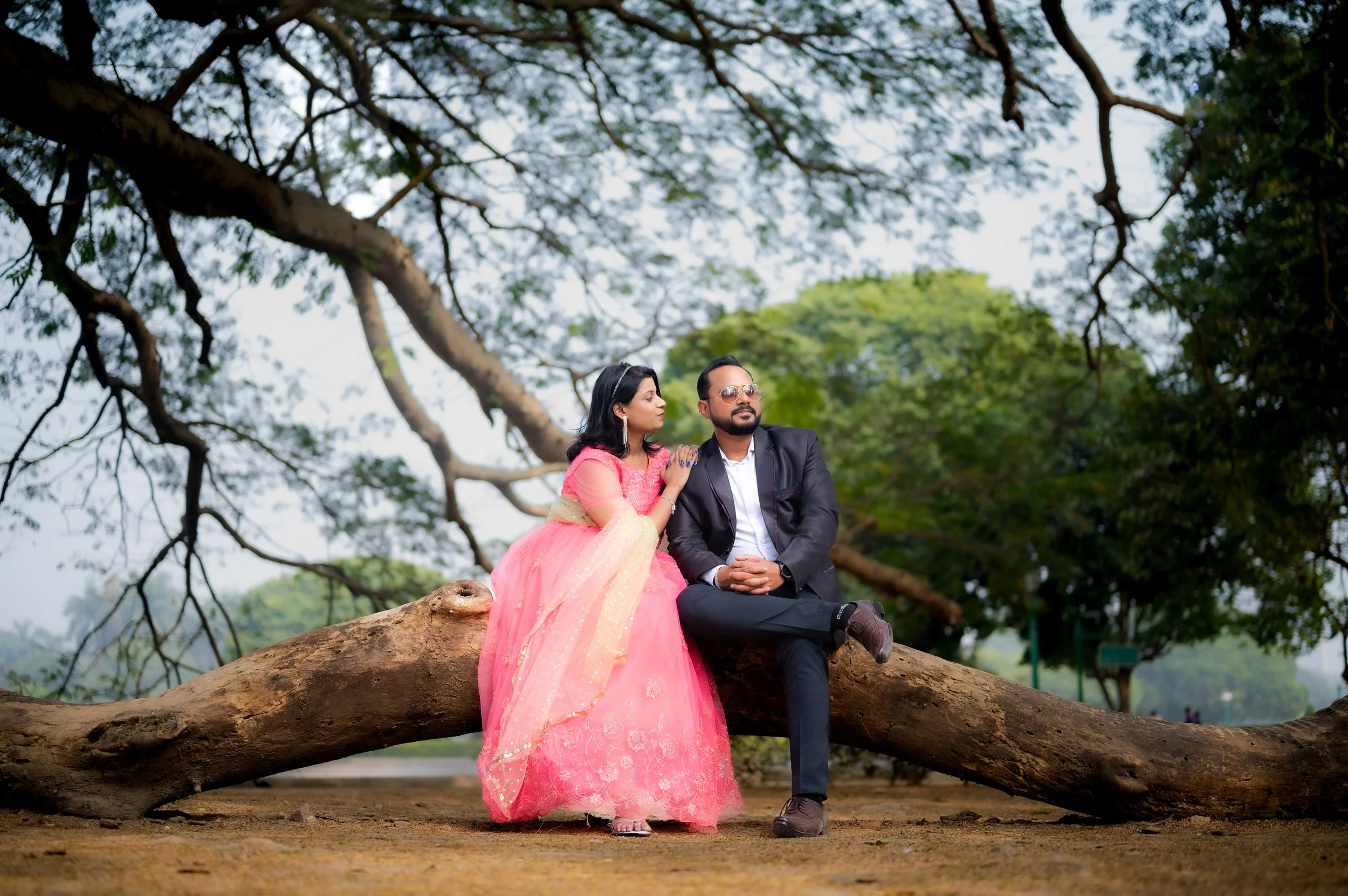 A man and woman sitting on a large tree branch outdoors. The woman is wearing a pink traditional dress with a dupatta, and the man is dressed in a formal black suit with sunglasses. They are surrounded by green trees, and the scene has a relaxing, natural setting.