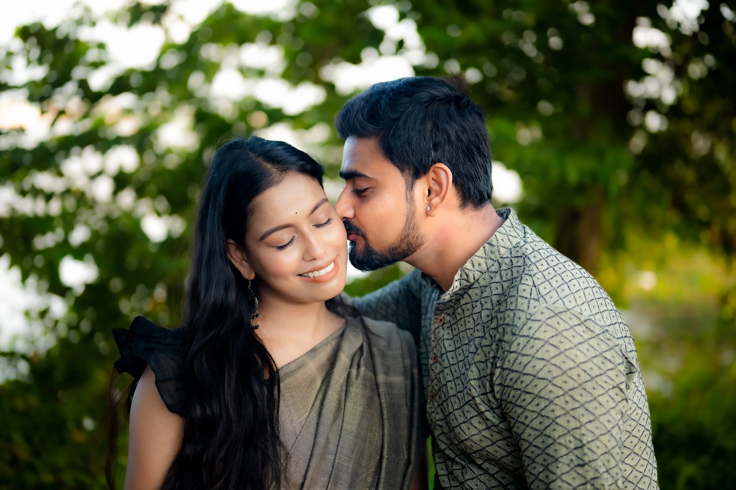 A man and woman share an intimate moment outdoors, with their faces close and eyes closed, surrounded by green trees.