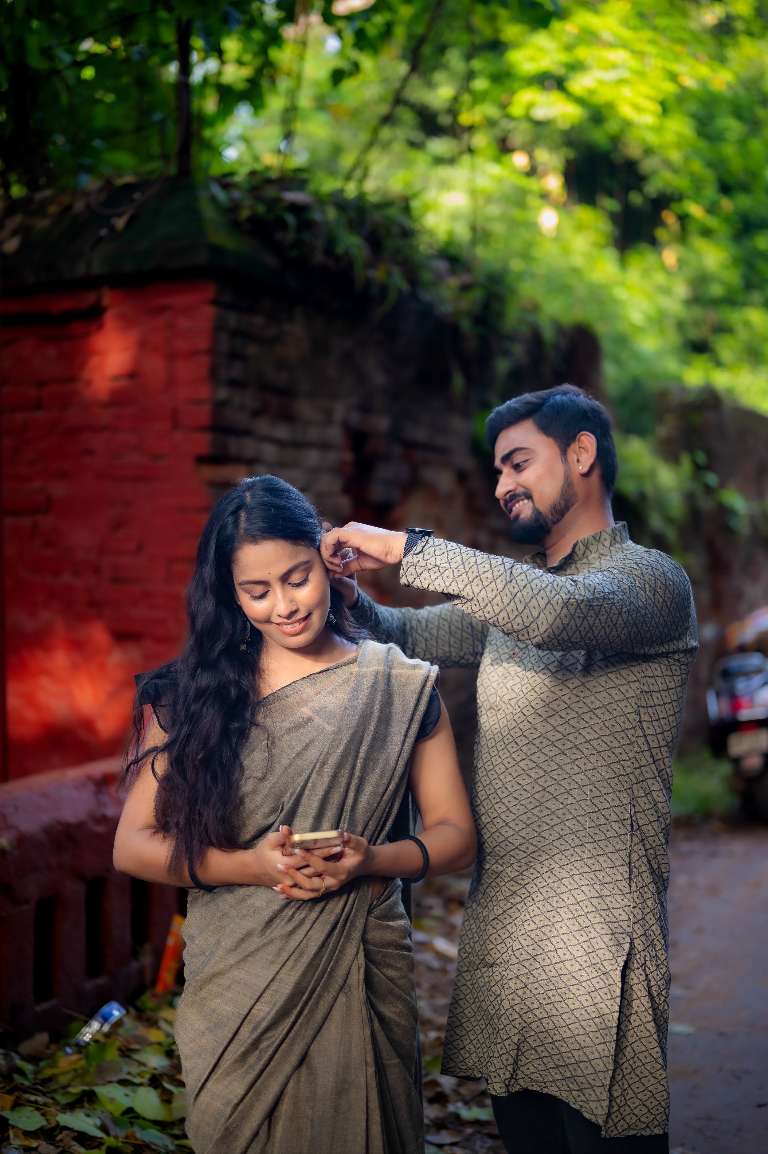 A man is styling a woman's hair outdoors in front of a red brick wall with green foliage, as she looks at her phone and smiles.