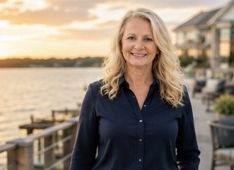 A smiling woman with blonde hair standing outdoors near a body of water during sunset, with houses in the background.