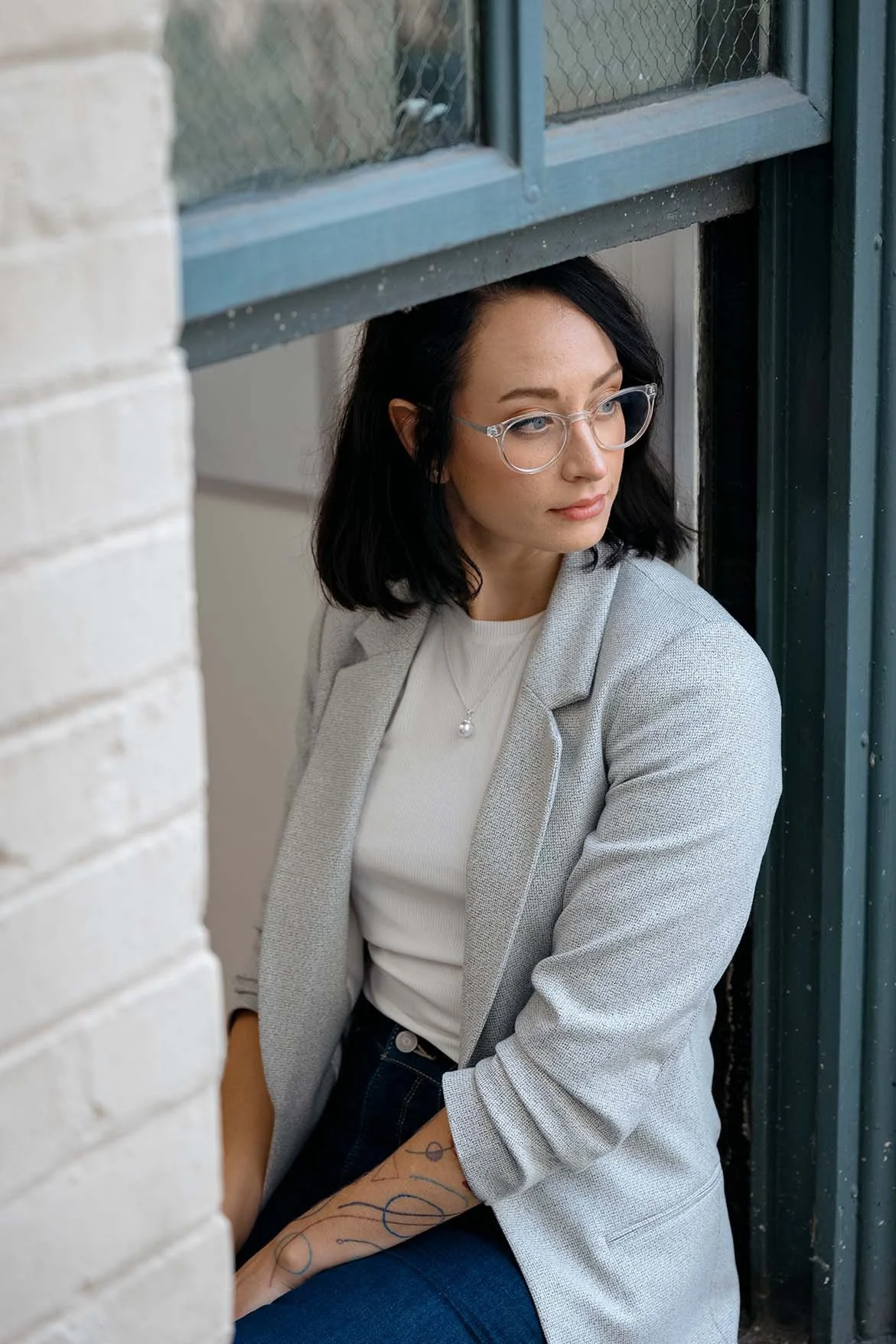A woman with black hair and glasses, wearing a grey blazer and a white top, sitting near a window and looking out, with a tattooed arm resting on her lap.