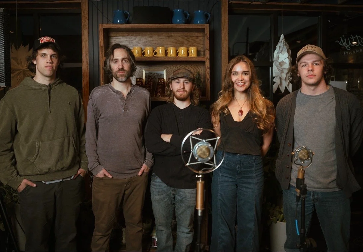 A group of five people standing indoors behind vintage microphones, posing for a photo in a cozy setting with wooden shelves and decorative items.