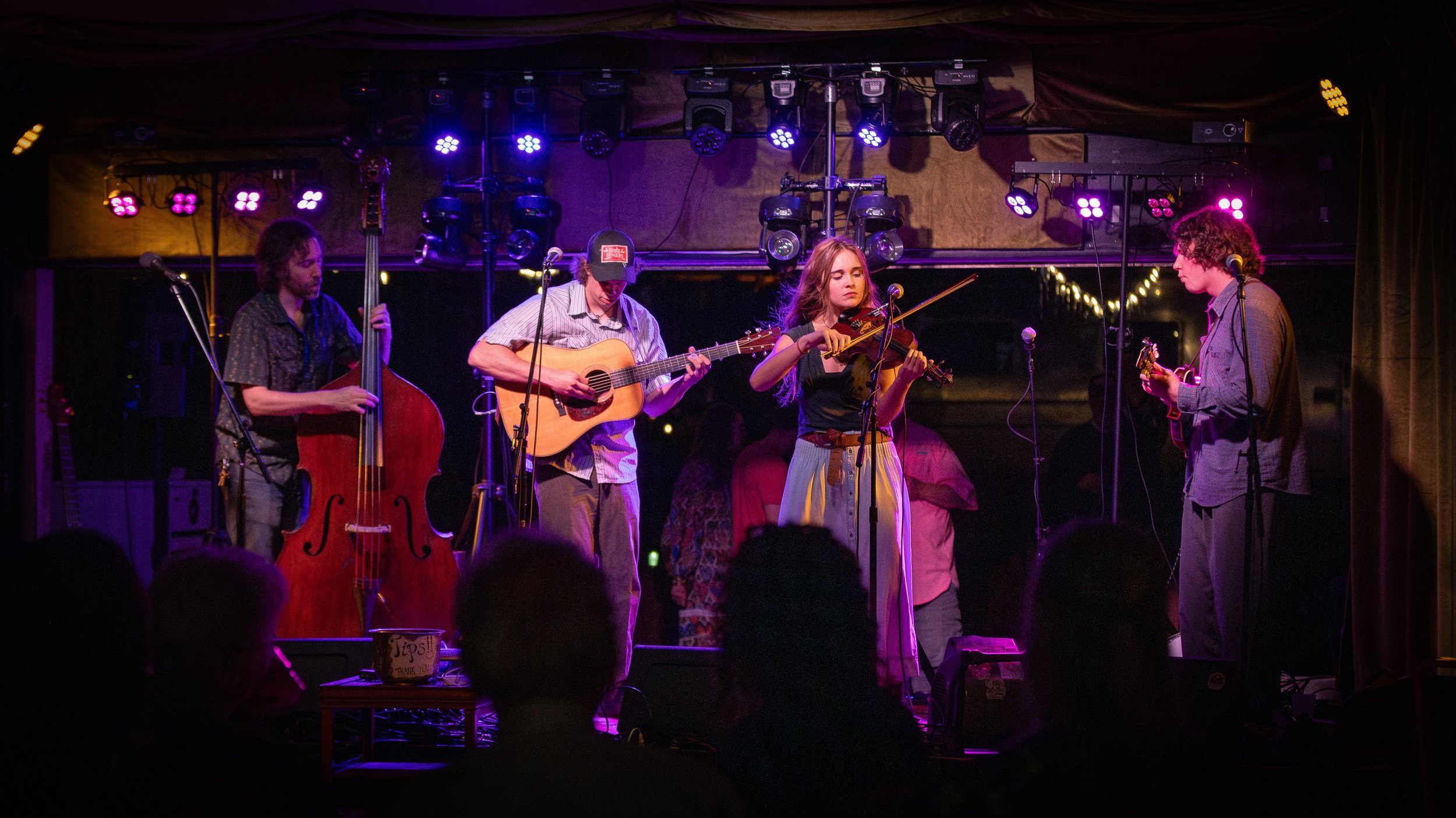 A group of musicians performing on stage with instruments including a double bass, guitar, and violin, under colorful stage lighting in a cozy venue.