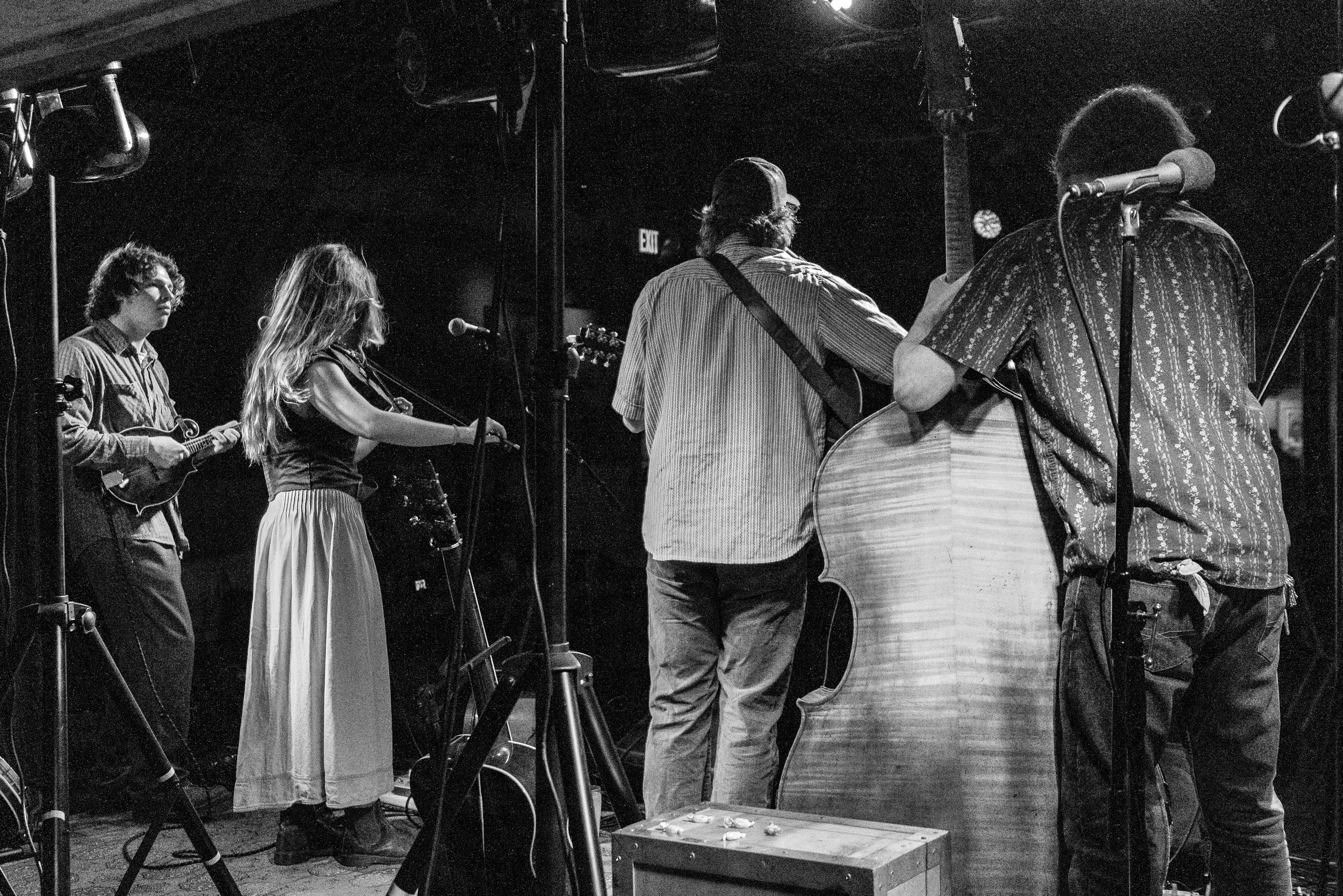 Four musicians performing on stage, three playing guitars and one singing into a microphone, in a dimly lit venue.