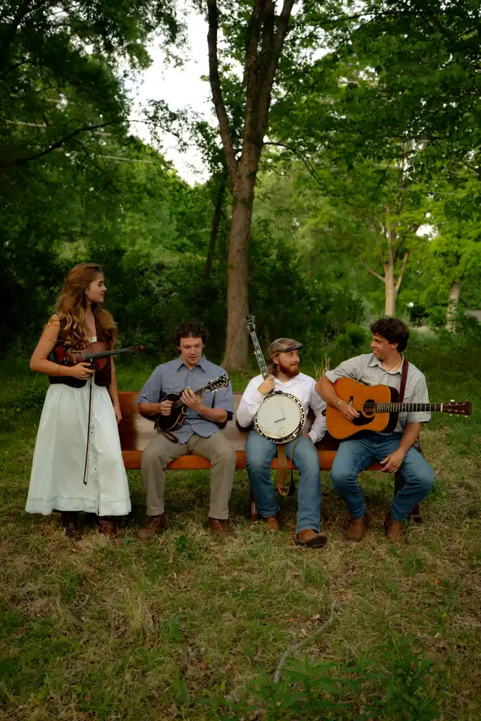 Four people playing musical instruments outdoors on a bench in a wooded area.