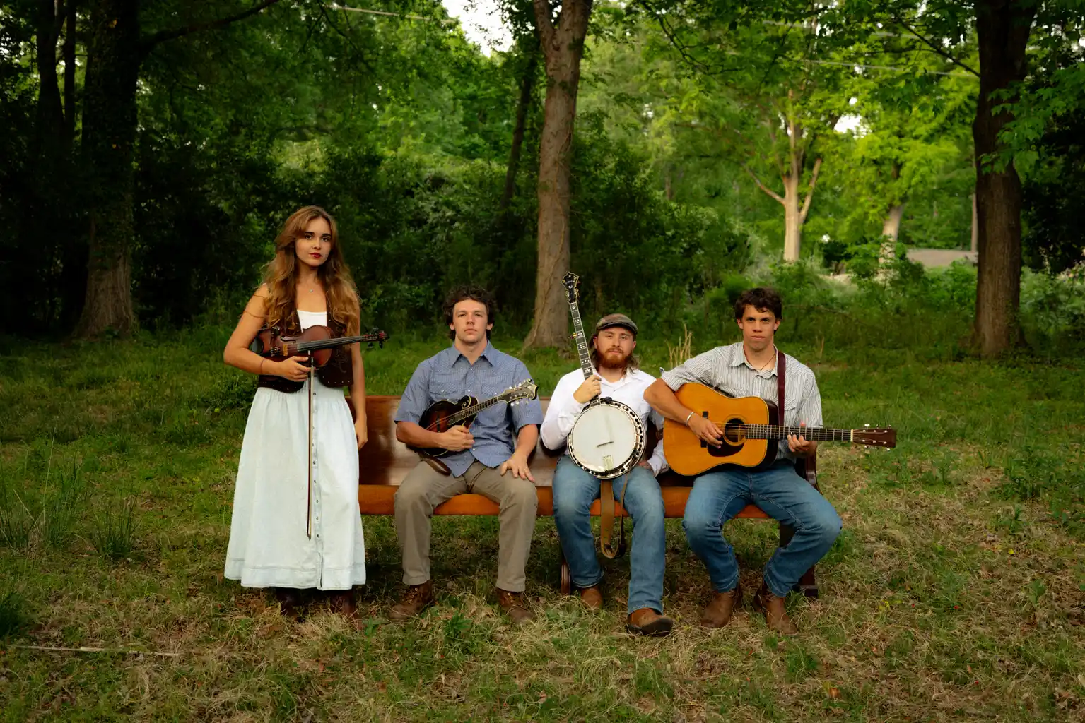 Four musicians playing instruments outdoors on a park bench in a lush green forest setting.