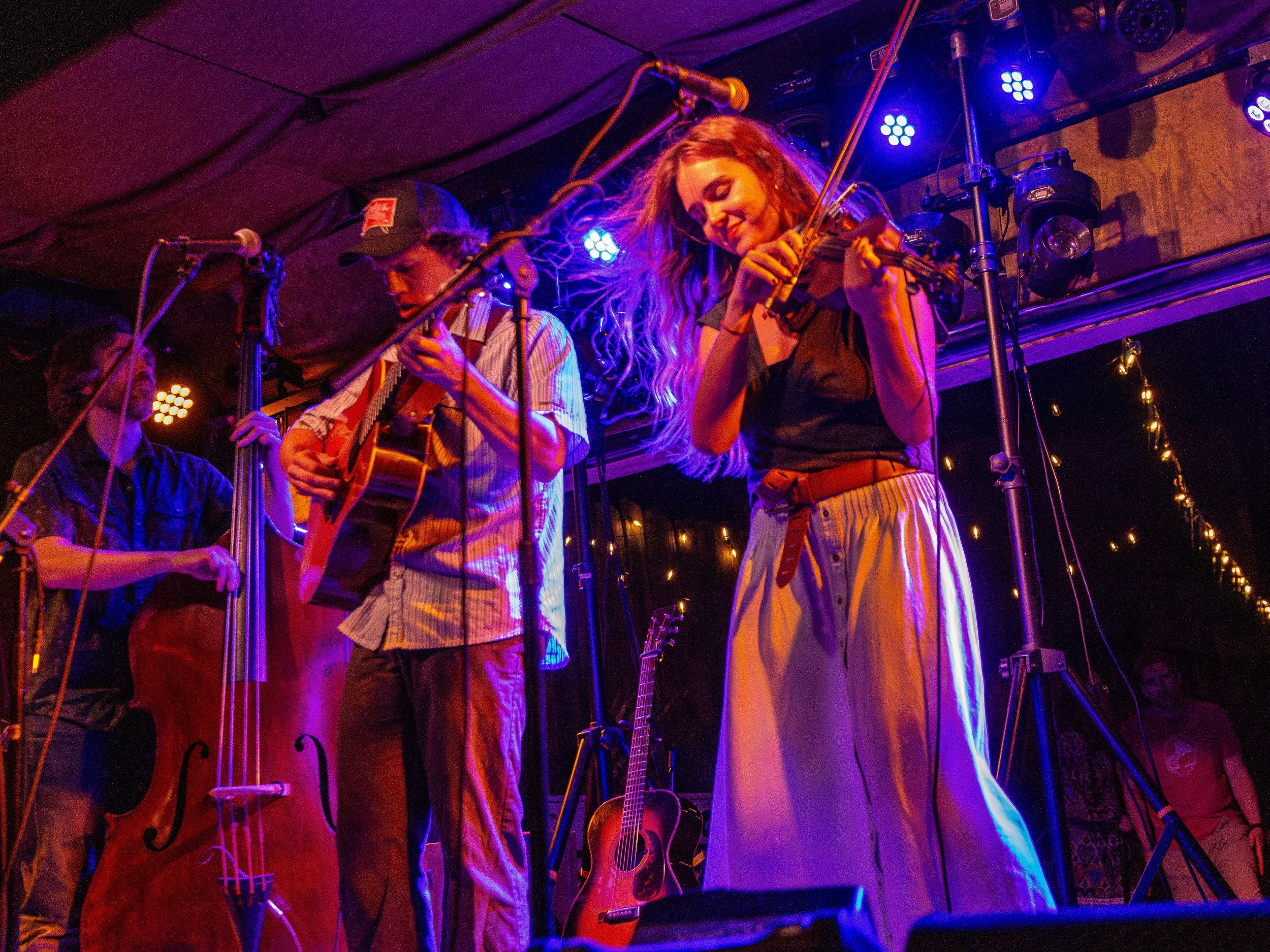 Musicians performing on stage with guitars and a violin inside a dimly lit venue with colorful lighting.