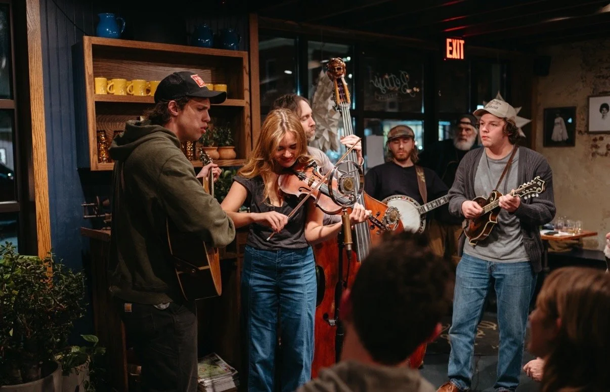A group of musicians playing instruments in a cozy indoor setting, with an audience in front of them.