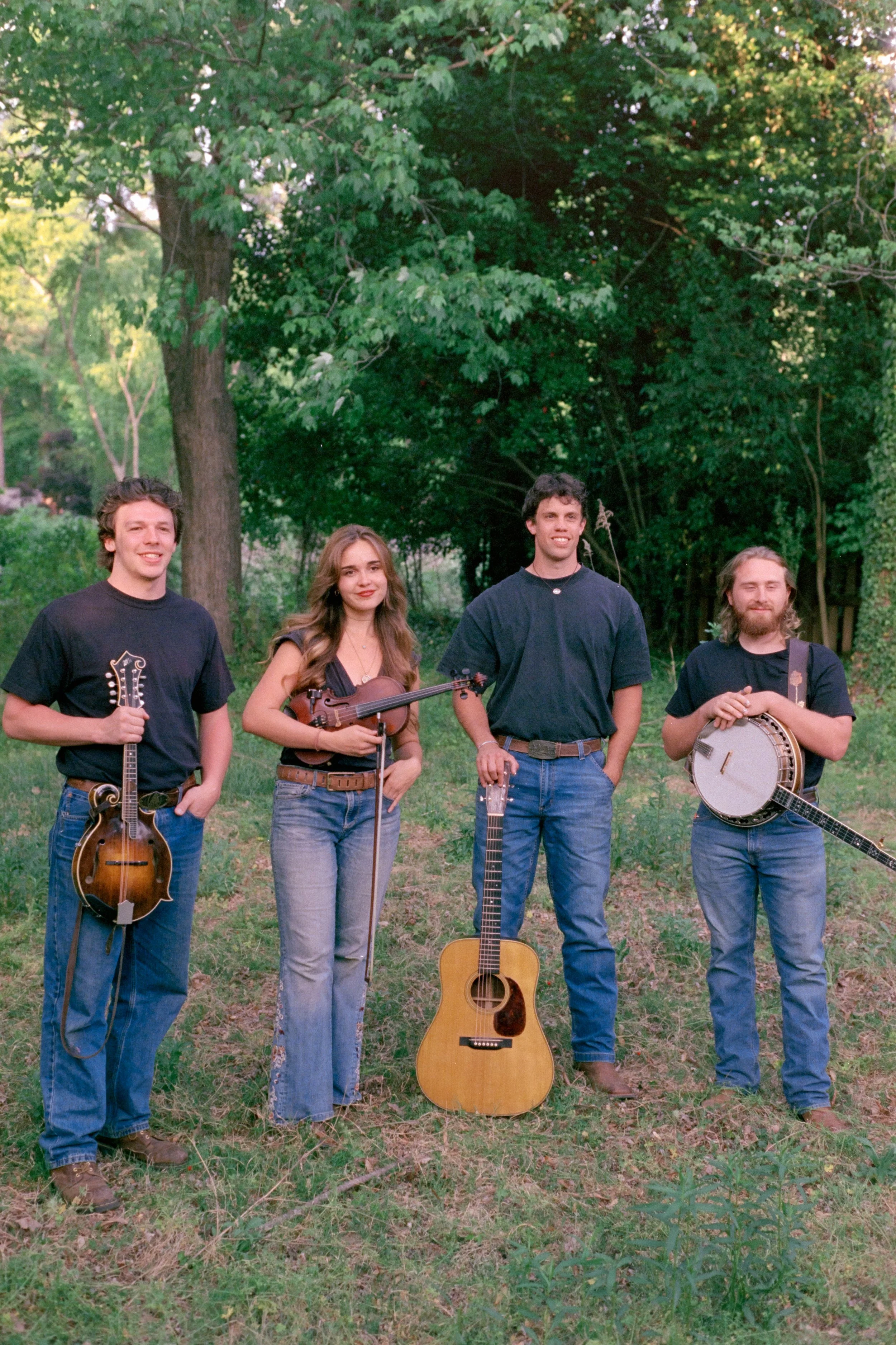 Four young adults standing outdoors on grass with trees in the background. They are holding musical instruments: a violin, a banjo, a guitar, and a tambourine.