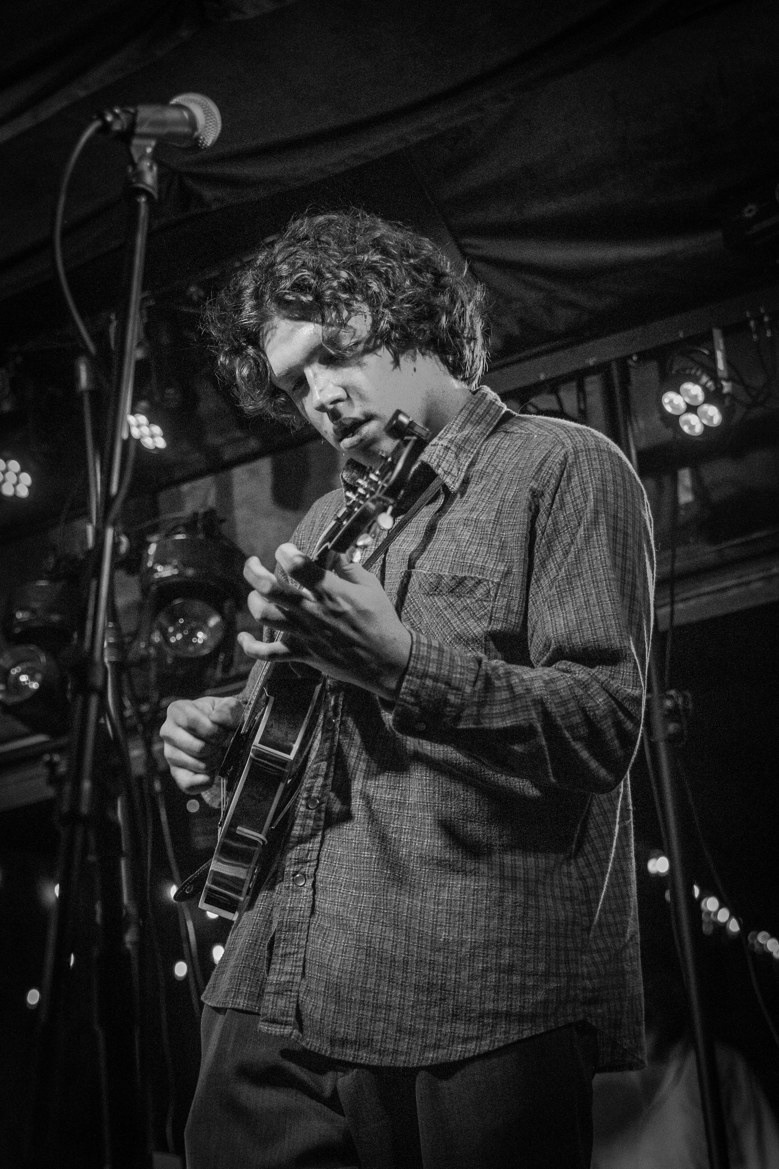 A young man with curly hair, wearing a plaid shirt, playing an acoustic guitar on stage with a microphone in front of him. The image is in black and white.