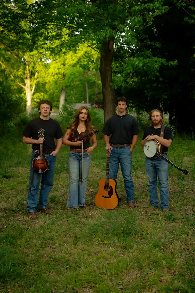 Four musicians holding instruments in a green park, standing under trees.