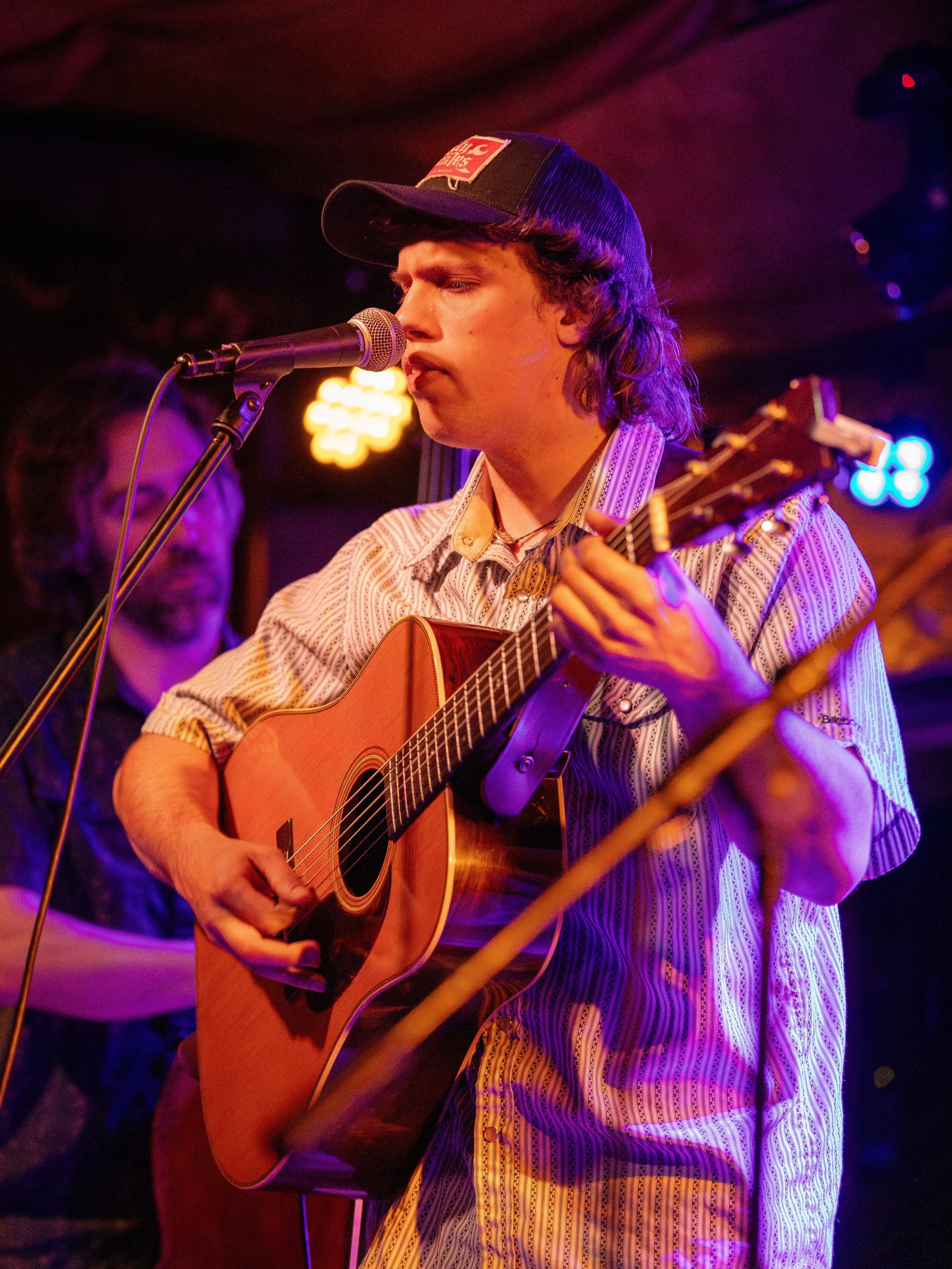A young man with long hair, wearing a trucker hat and a patterned shirt, is playing an acoustic guitar and singing into a microphone on a dimly lit stage with colorful lights.