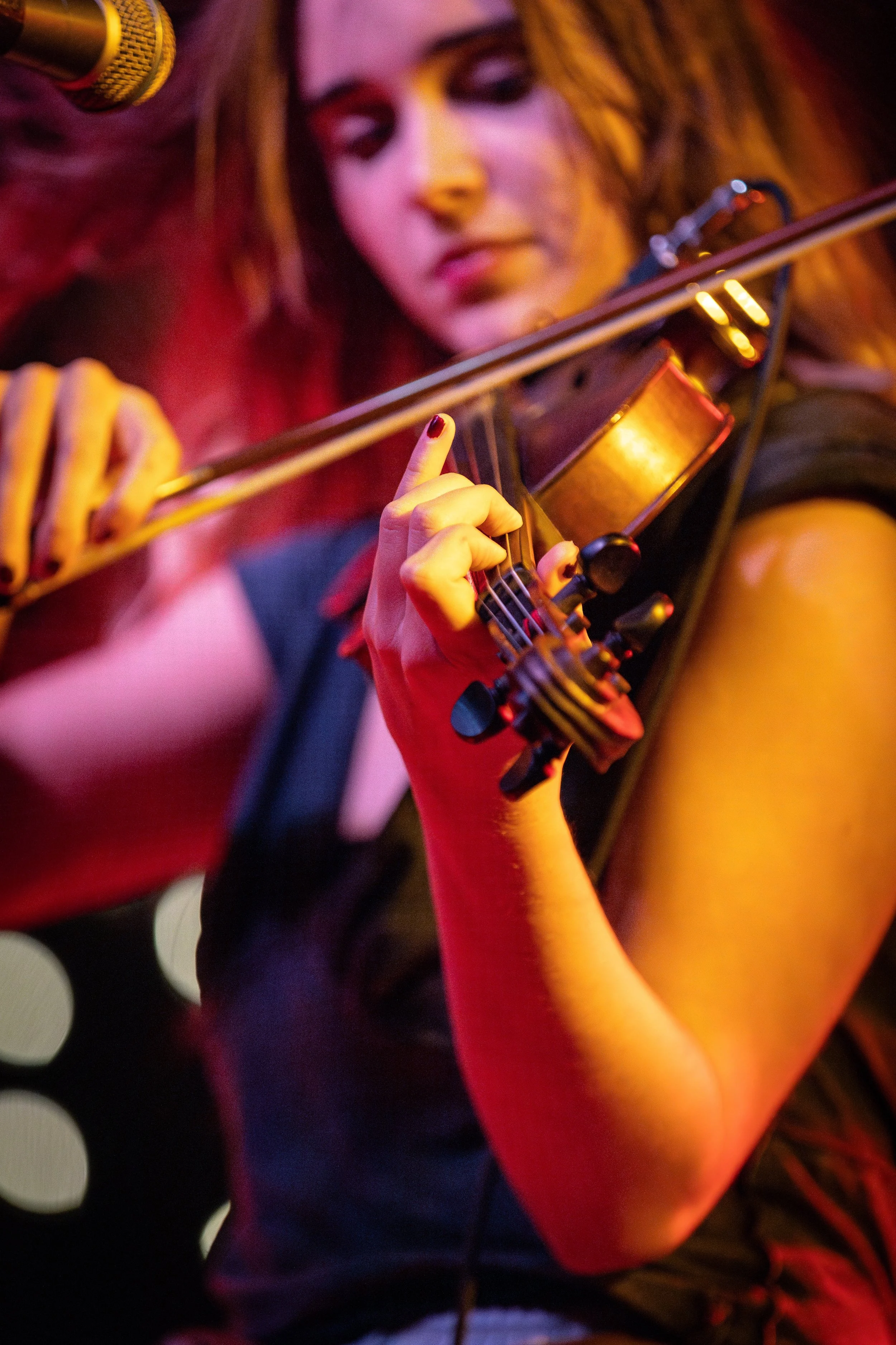 A woman playing the violin on stage, with a microphone nearby and soft, colorful lighting.