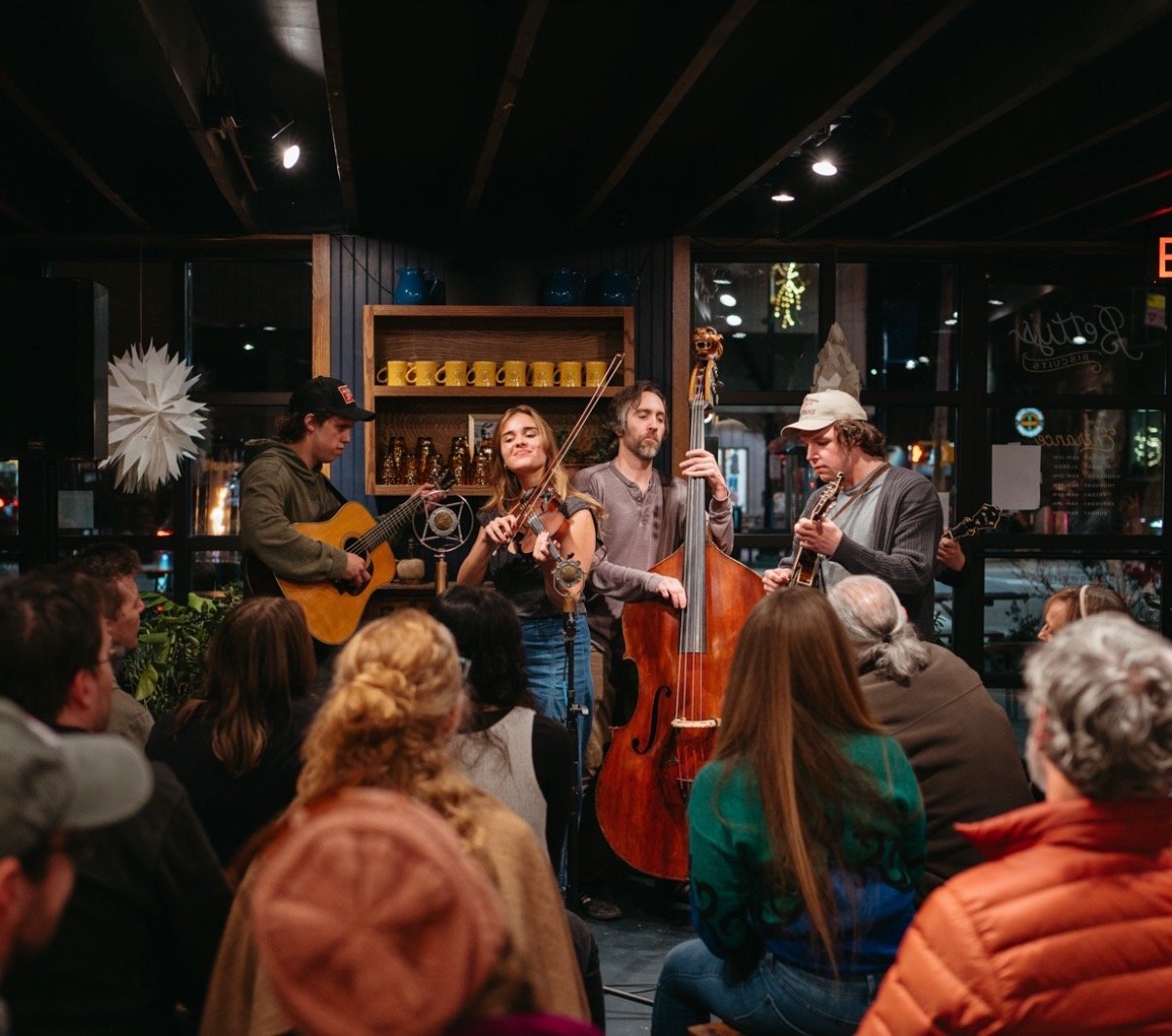 A band of four musicians performing live at a cozy indoor venue while an audience watches, with the background featuring glass windows and Christmas decorations.