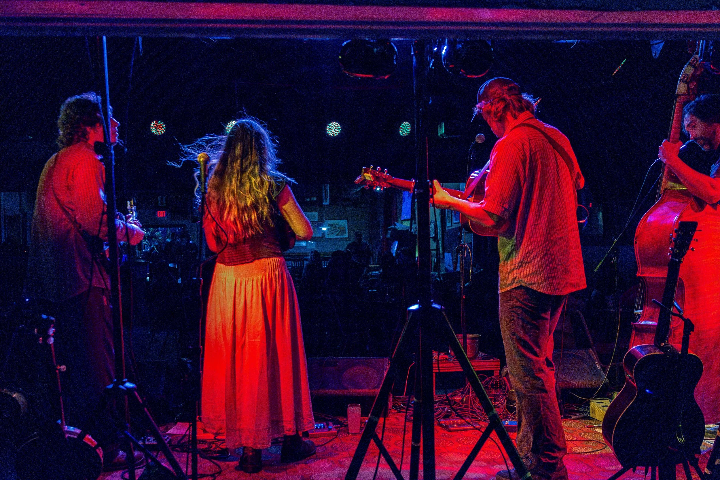 A live music performance in a dimly lit venue featuring four musicians, two women and two men, playing guitars and singing on stage with microphone stands and colorful stage lighting.