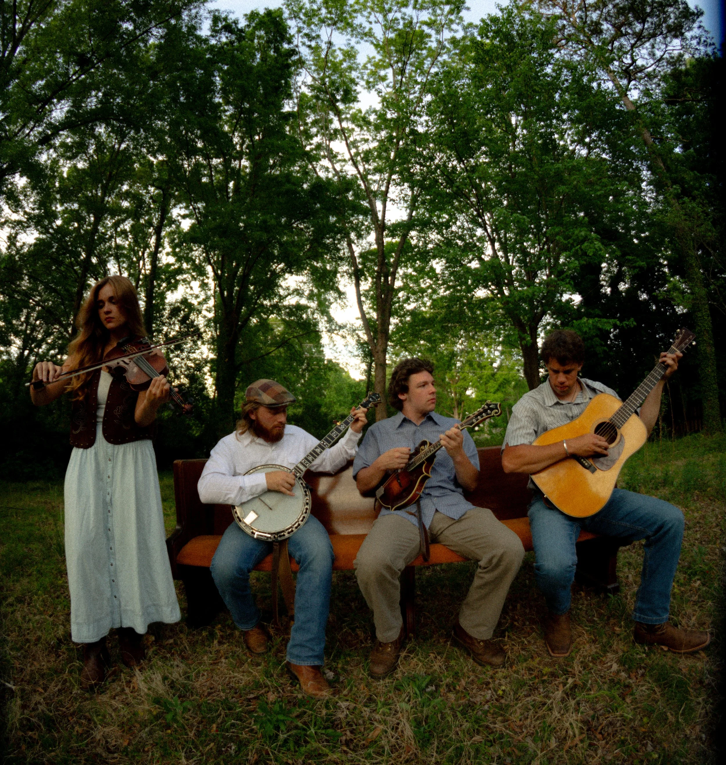 A group of young people, playing guitars and a violin, sitting and standing outdoors among green trees on a cloudy day.