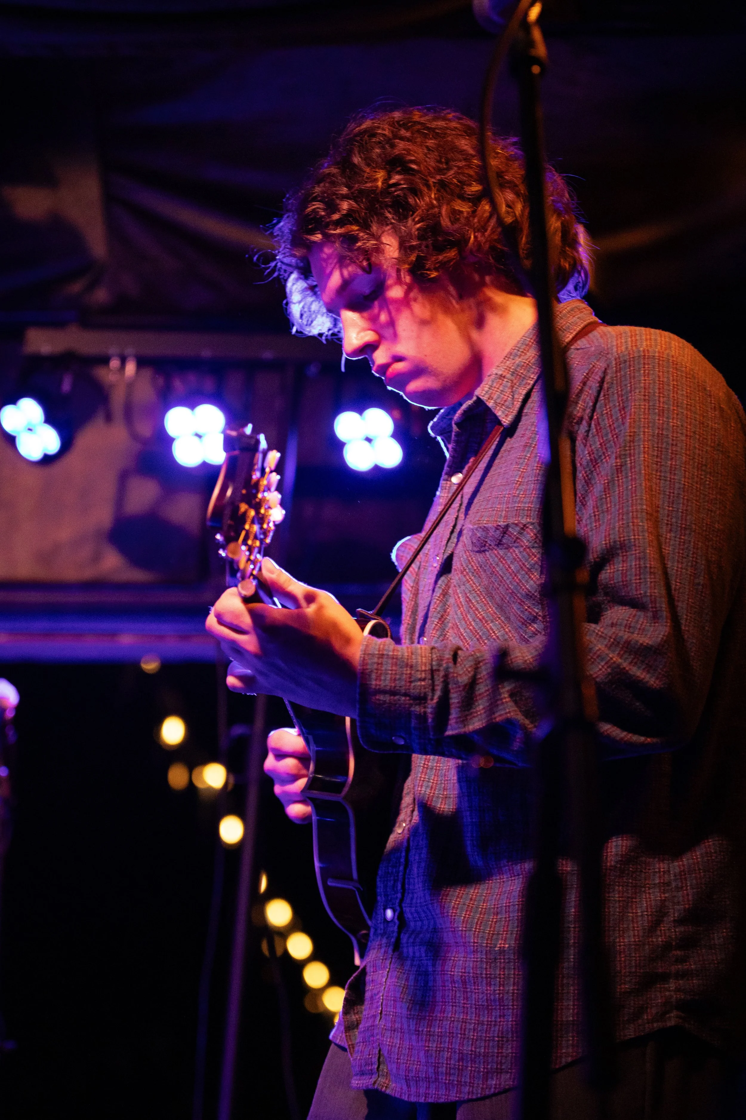 A musician with curly hair wearing a plaid shirt playing an electric guitar on stage, illuminated by colorful purple and blue lights, with blurred stage lights in the background.