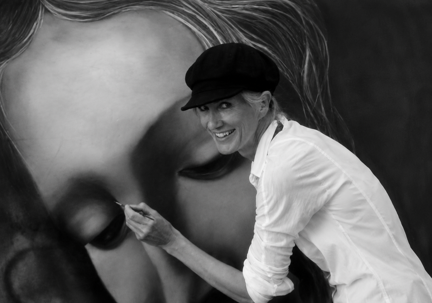 A woman smiling and wearing a cap, painting a large black and white portrait of a person's face.