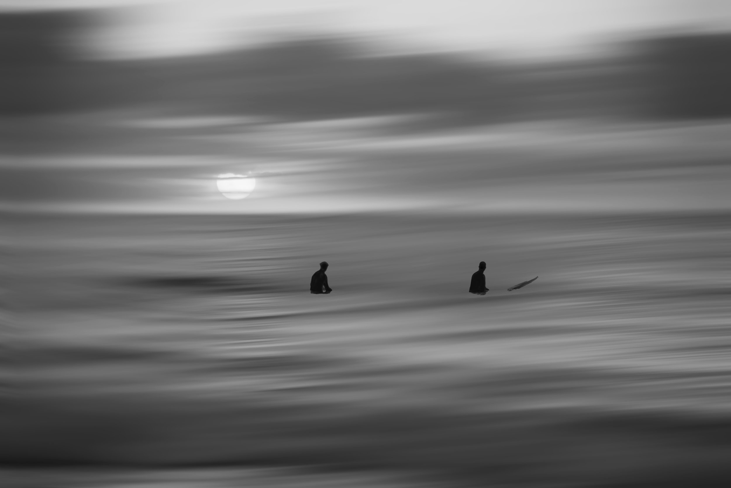 A black and white photo of two surfers in the ocean, one sitting and one lying on a surfboard, with the sun visible through cloudy sky.