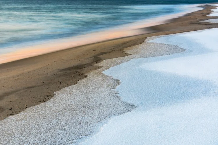 Ocean waves washing over sandy beach with snow and ice along the shoreline at sunset.