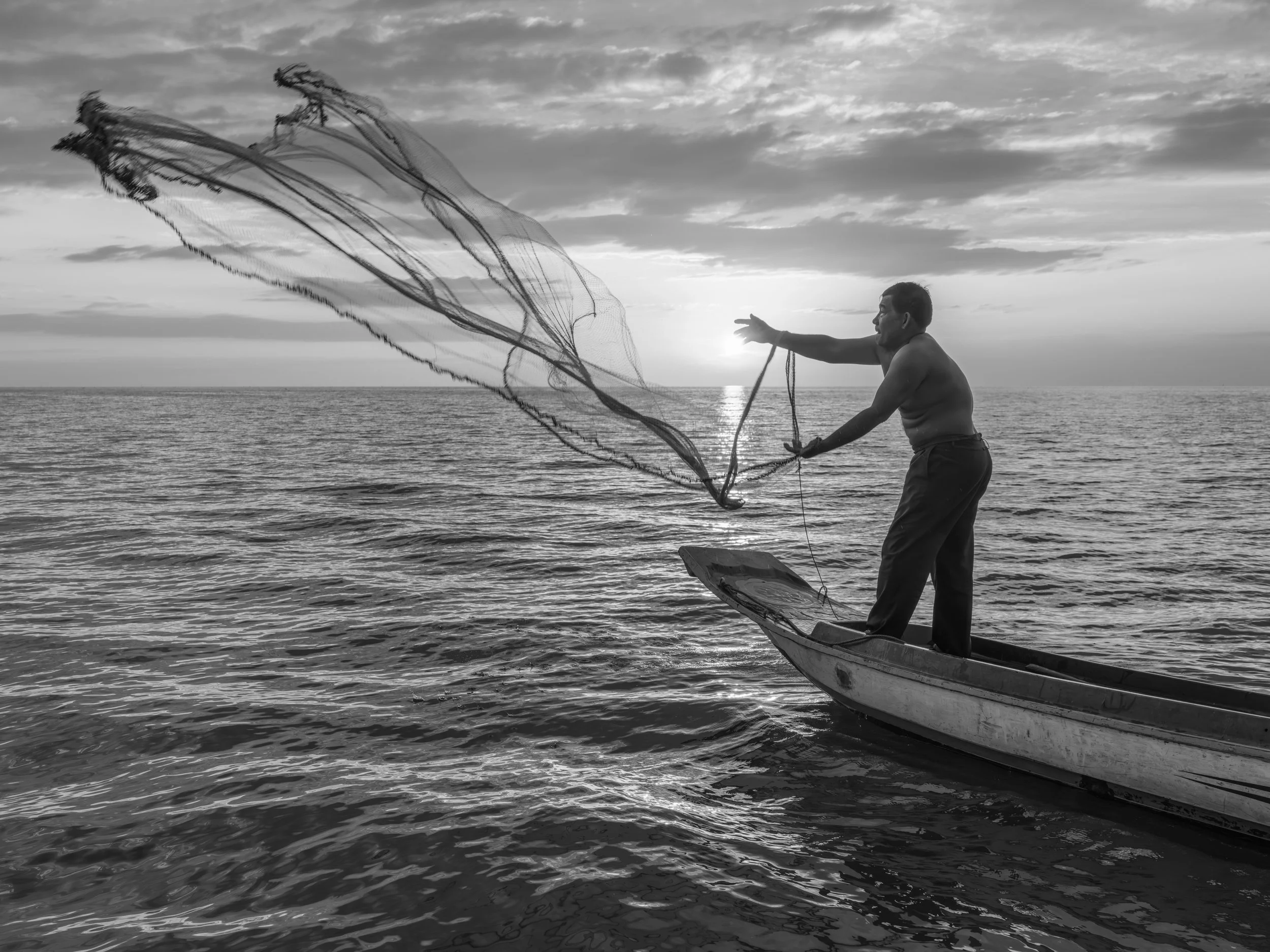 A man standing on a small boat in the water, tossing a fishing net while the sun sets or rises over the ocean.