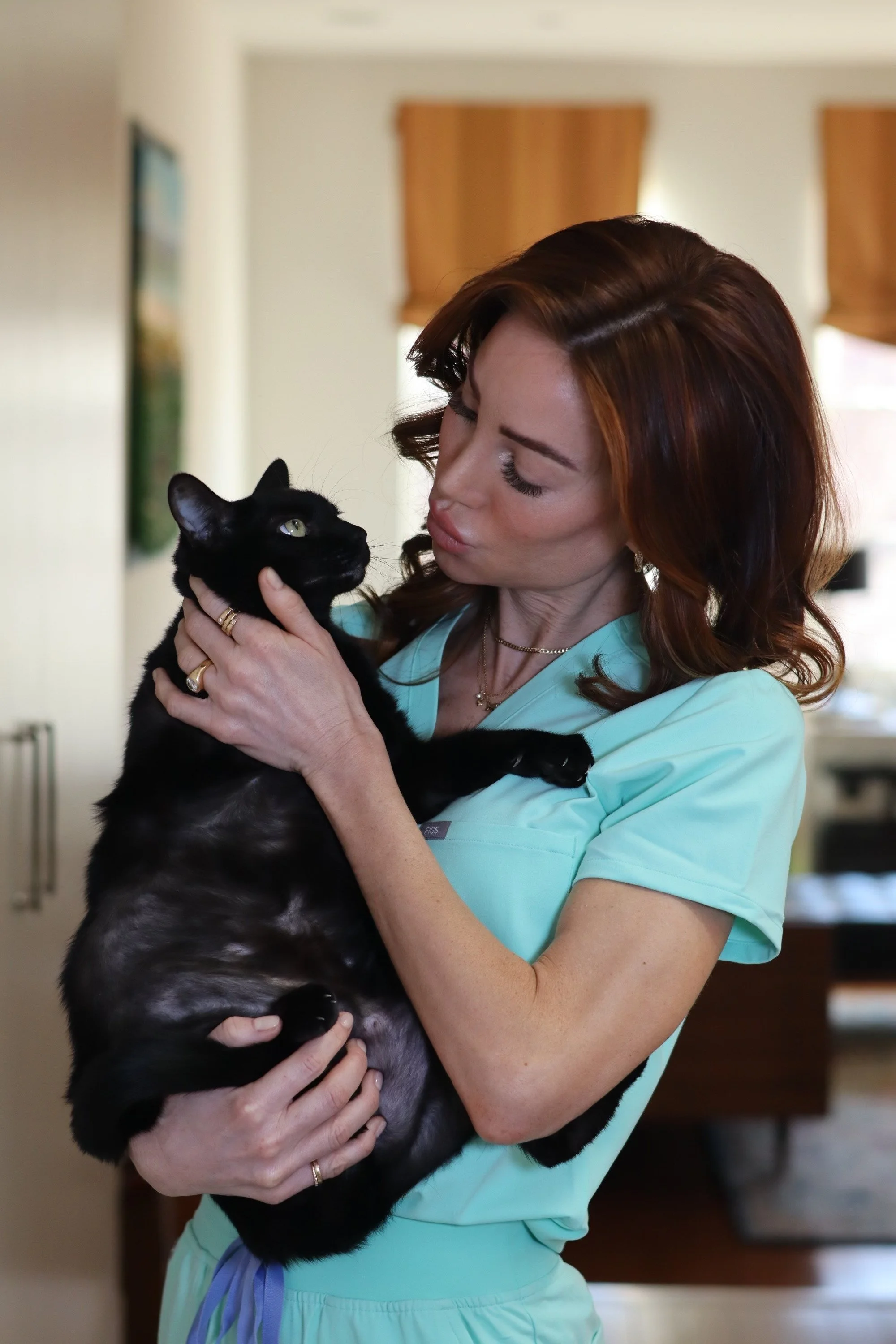 A woman in medical scrubs holding a black cat with green eyes indoors.