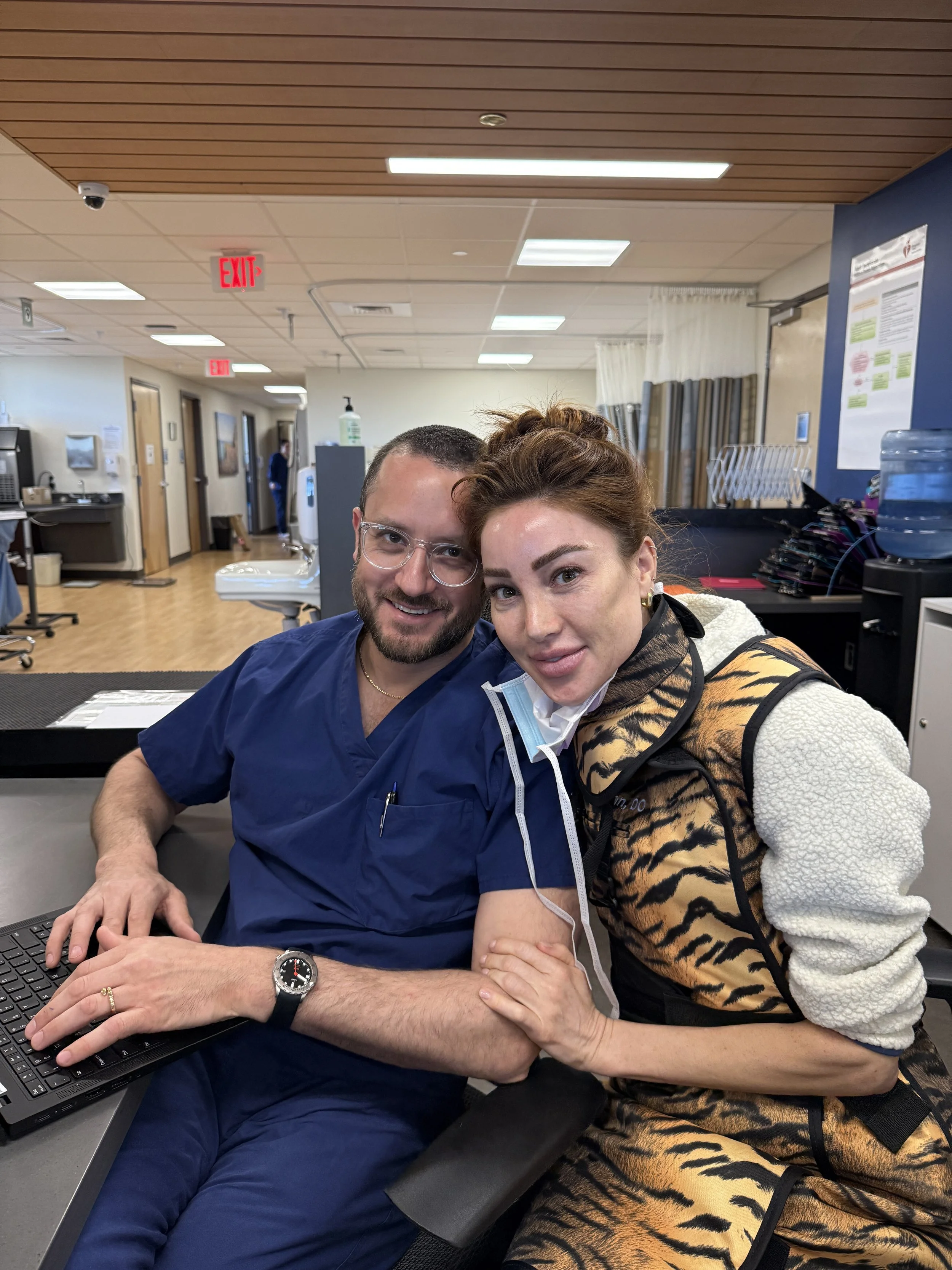 Two people sitting at a desk in a medical setting, smiling and looking at the camera. The man is wearing blue medical scrubs and glasses, and the woman has her face partially covered with a mask pulled down, wearing a tan and black tiger-striped hoodie. Background includes medical equipment, water dispenser, informational posters, and an exit sign.