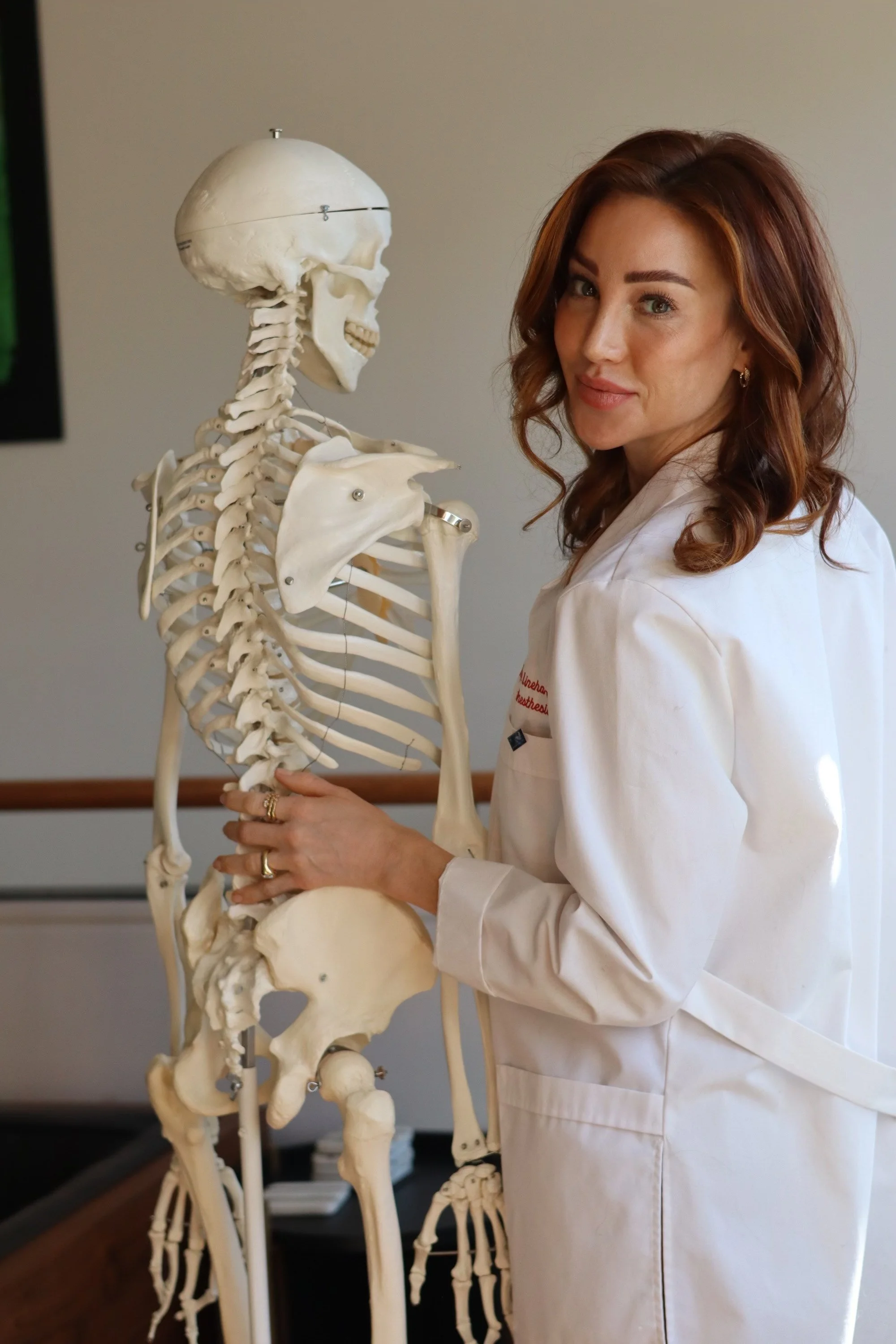 A woman in a white lab coat standing next to a human skeleton model.
