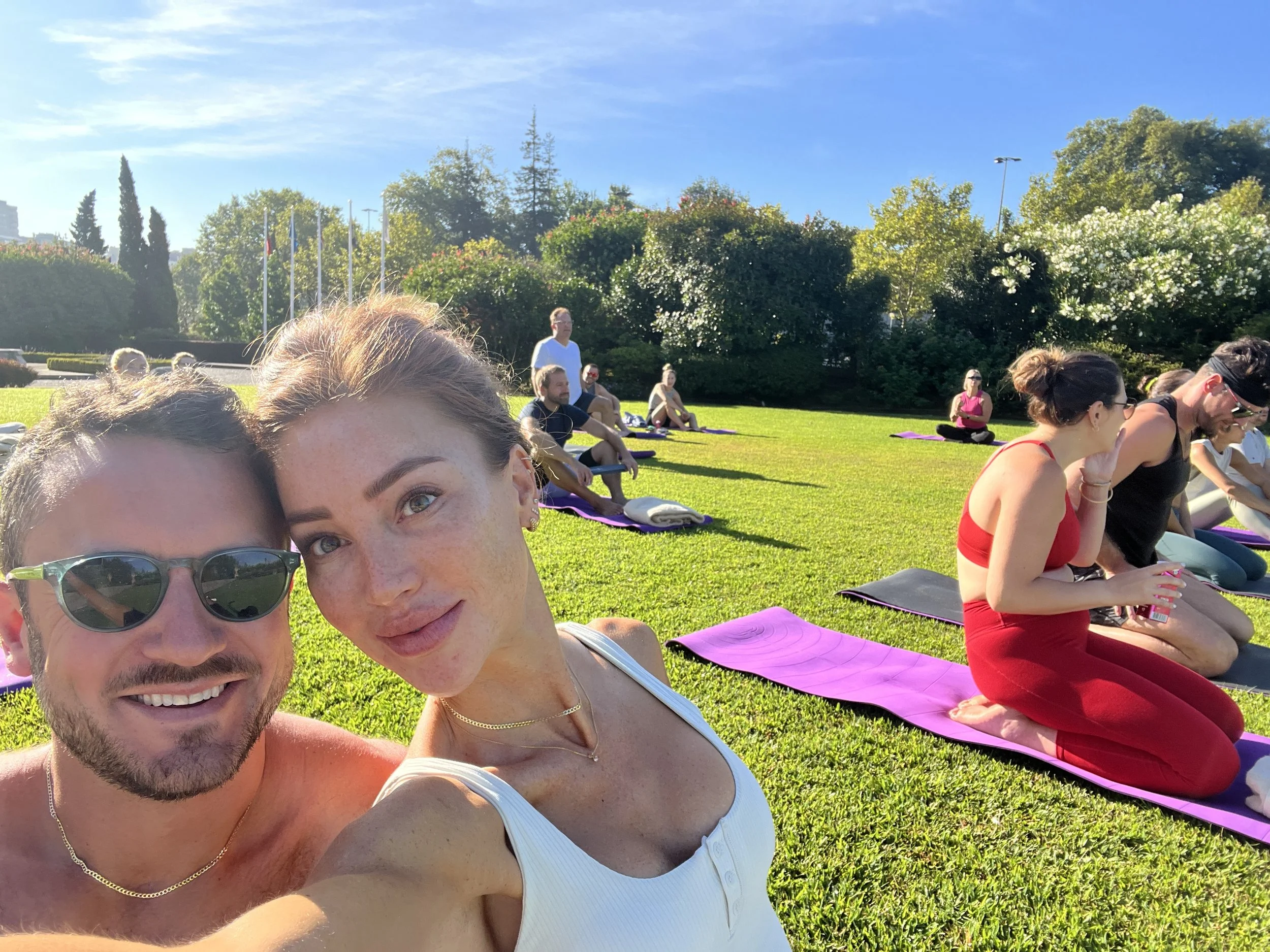 A group of people practicing yoga outdoors on purple mats in a park with green grass and trees on a sunny day. Two people in the foreground are taking a selfie, smiling at the camera.