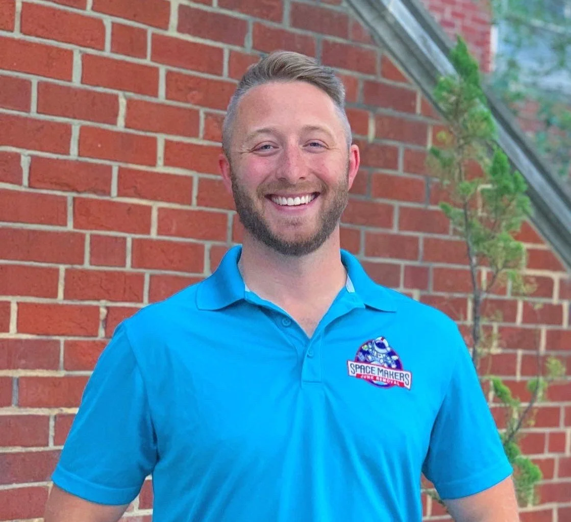 A smiling man with a beard and short hair wearing a blue polo shirt who works for Traction Local SEO because he is an SEO Expert, standing outside in front of a red brick wall and some greenery.