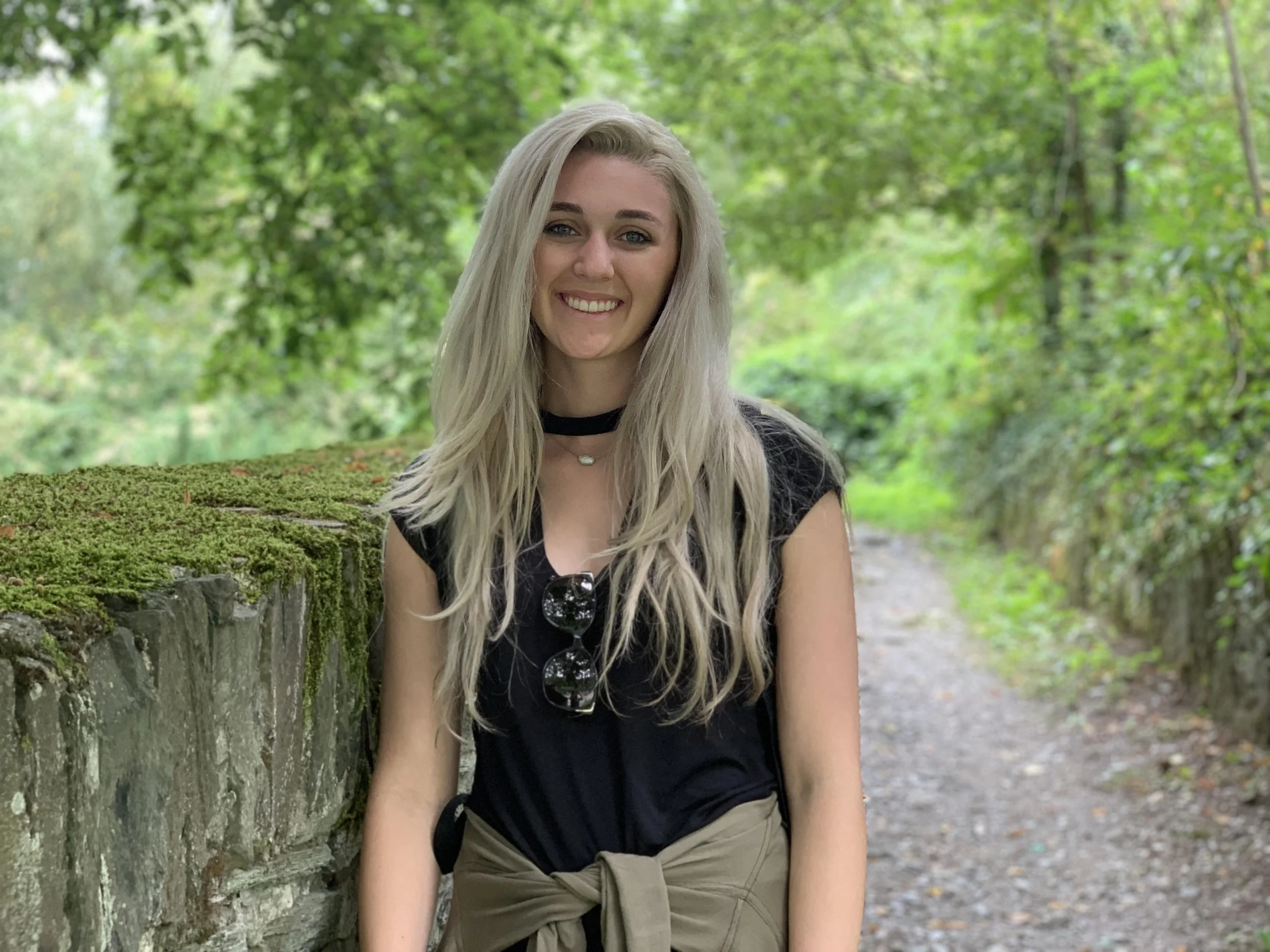 A smiling young woman with long blonde hair standing outdoors in front of a mossy stone wall and green trees, wearing a black top, beige shorts, a black choker, and sunglasses hanging from her shirt, ready to work on improving SEO for local business