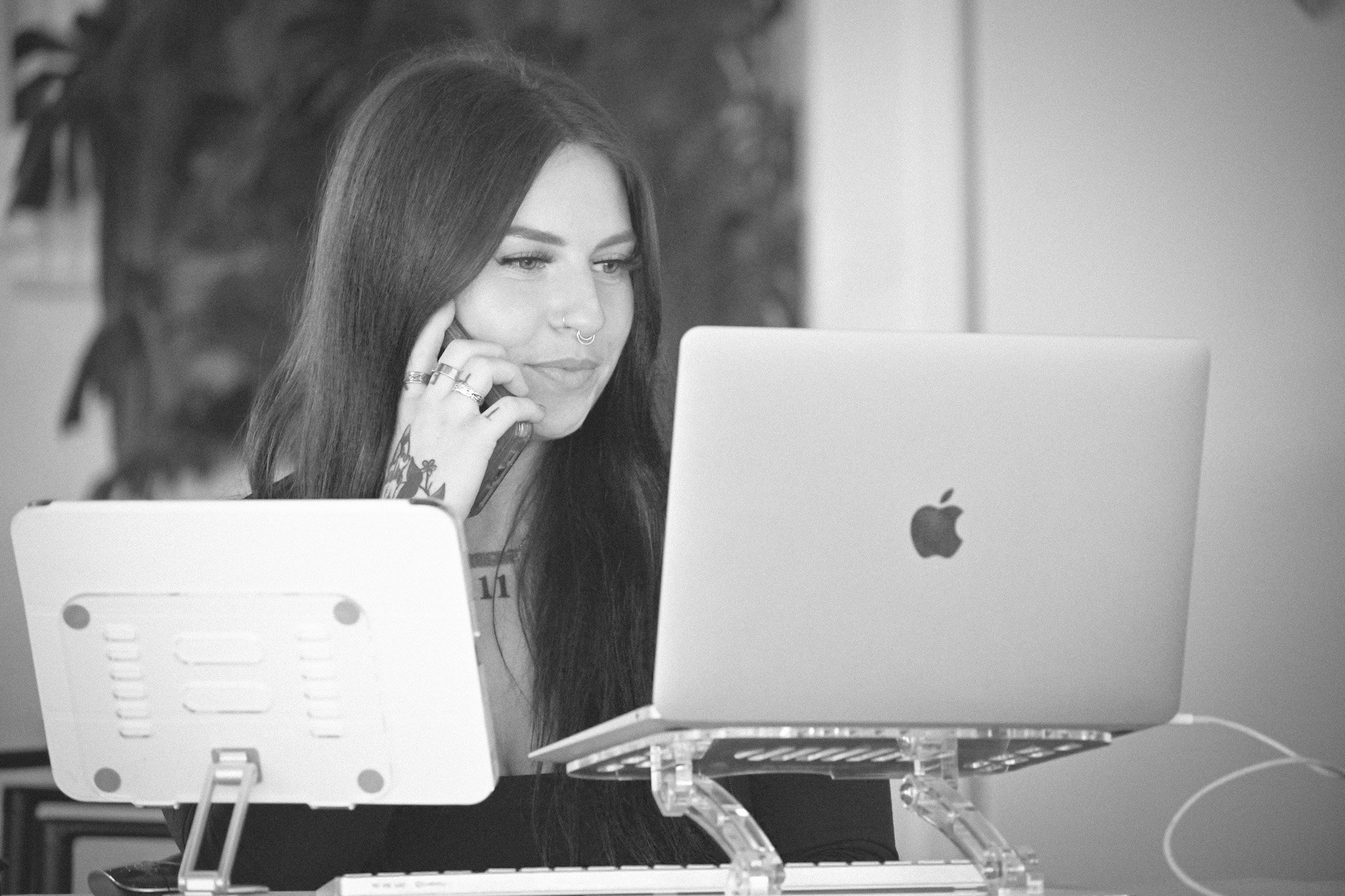 A woman with dark hair and tattoos on her arm, wearing rings and a nose ring, looks at her laptop. She is seated at a desk with a white Apple computer and a stand, with another device in front of her. The background shows a blurred wall with a framed picture.