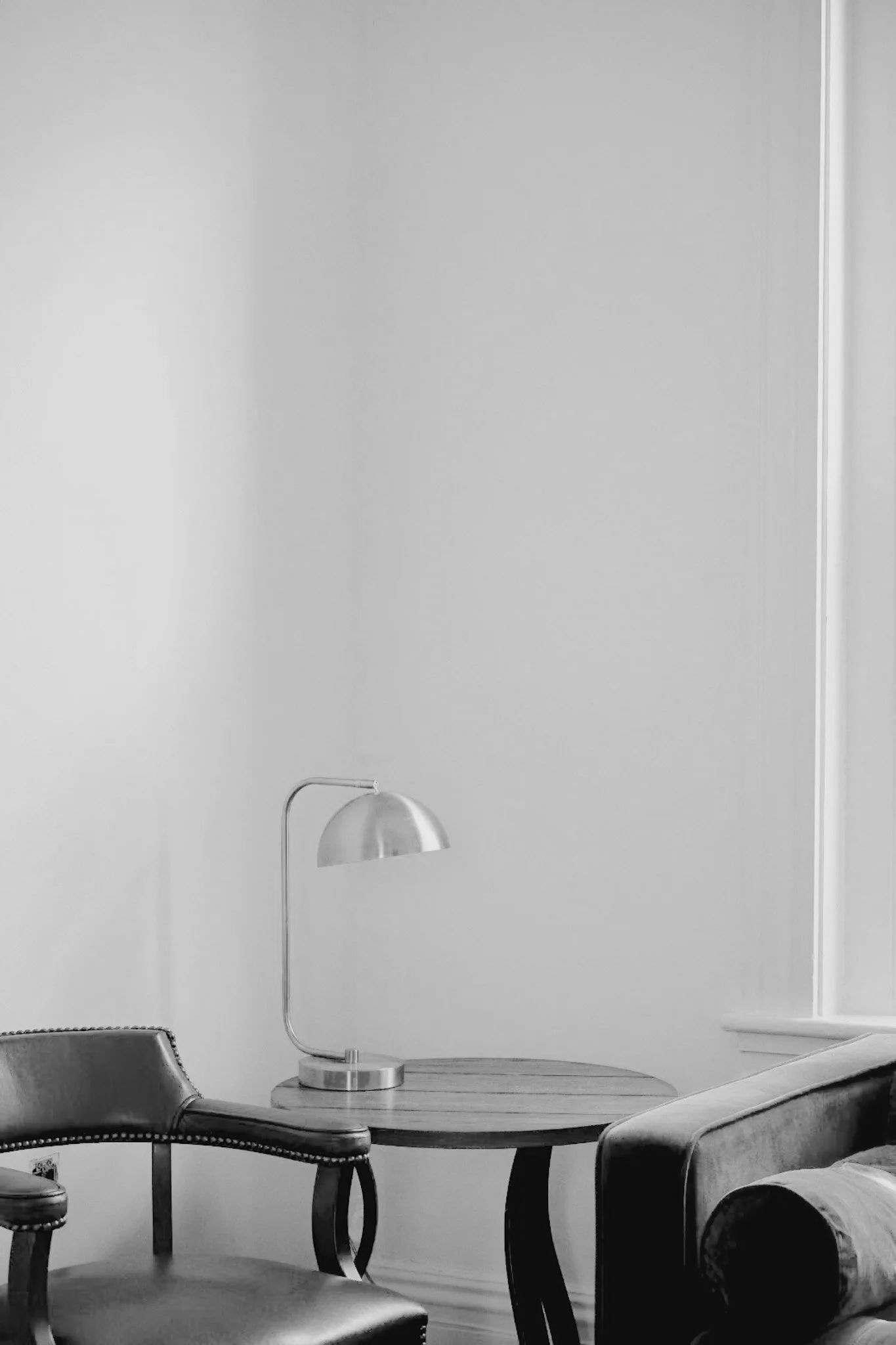 Black leather chairs around a round wooden table with a modern metallic desk lamp on it, positioned in a room with plain white walls.