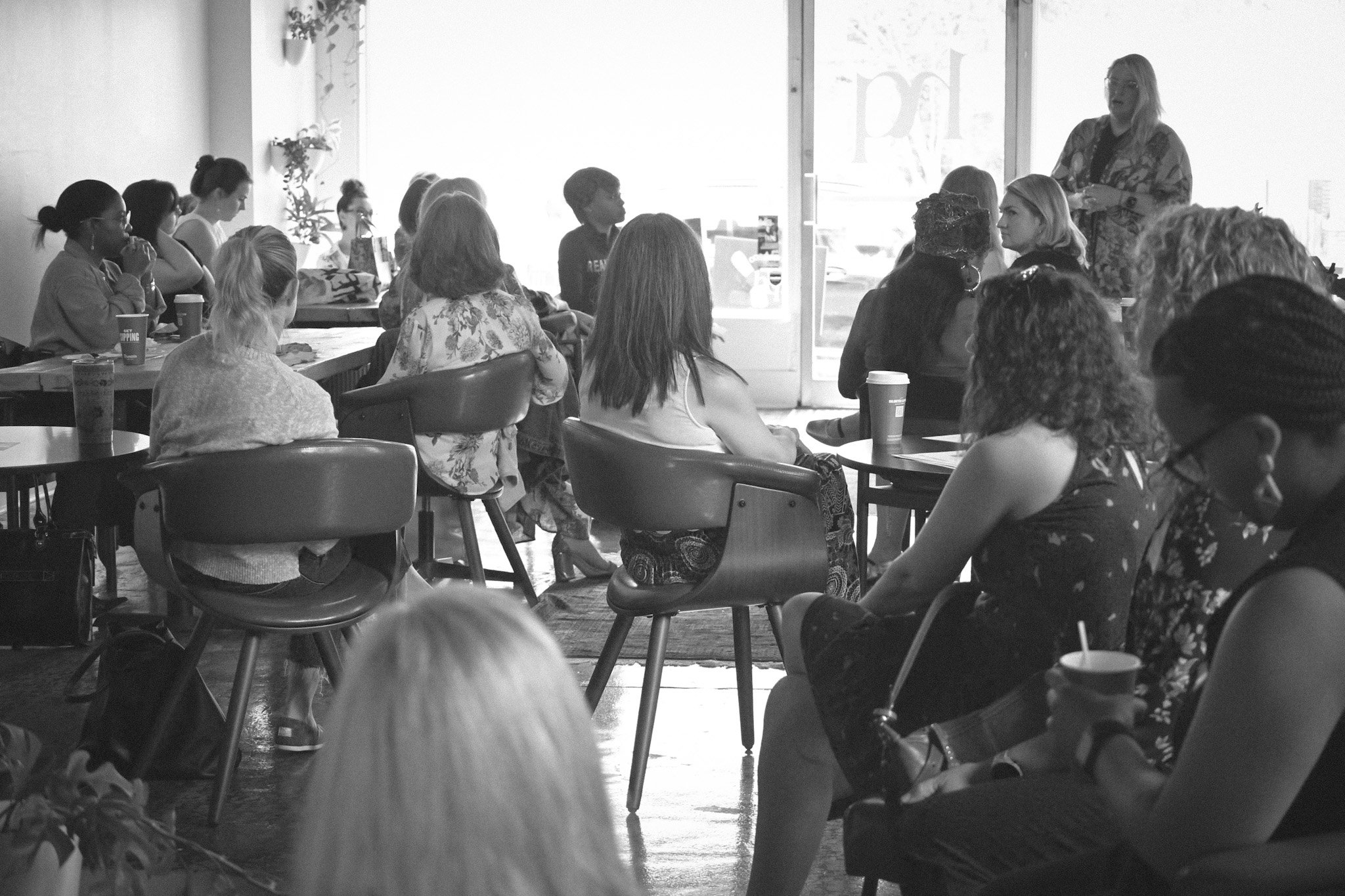 A woman appears to be giving a presentation or speech to a group of diverse people seated in a cafe or coffee shop with large windows letting in natural light. The audience is attentive, with some taking notes or enjoying drinks.