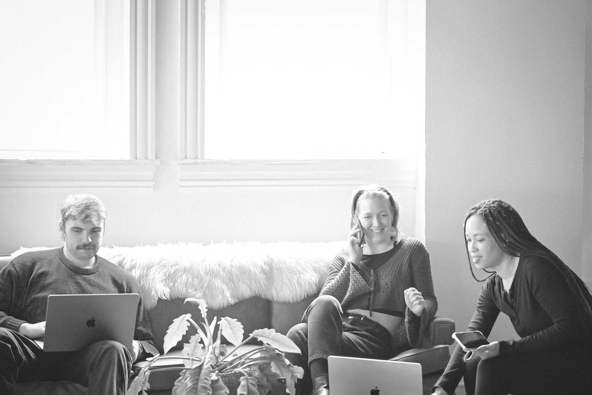 Three people sitting together in a cozy room, with a man using a laptop, a woman talking on the phone, and another woman looking at her phone, with a window and plants in the background.