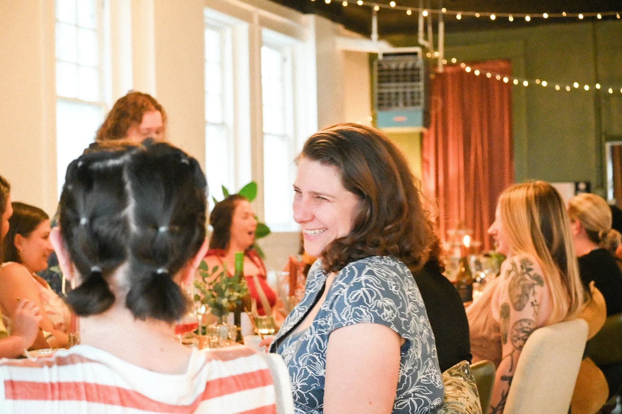 People gathered at a social event, smiling and engaging in conversation, with decor including string lights and large windows.