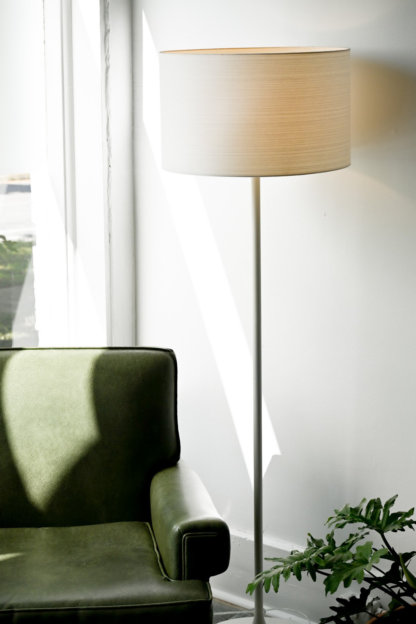 A green armchair next to a tall white floor lamp with a beige lampshade and a potted plant with green leaves near a sunlit window.