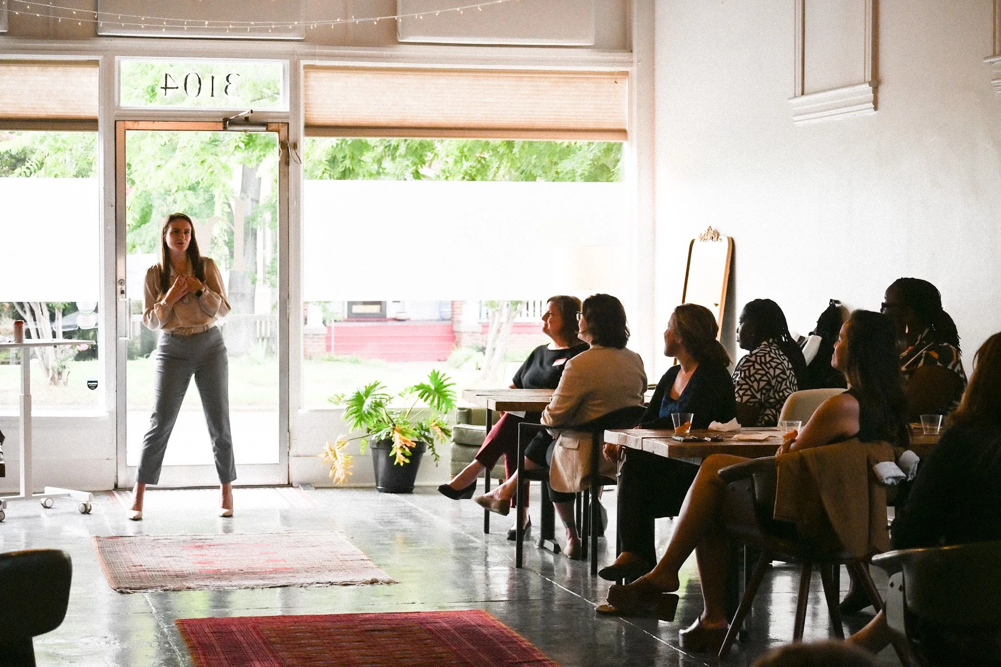 A woman standing and speaking in front of an audience during a presentation inside a well-lit room with large windows and hanging string lights. The audience is seated and attentively listening.
