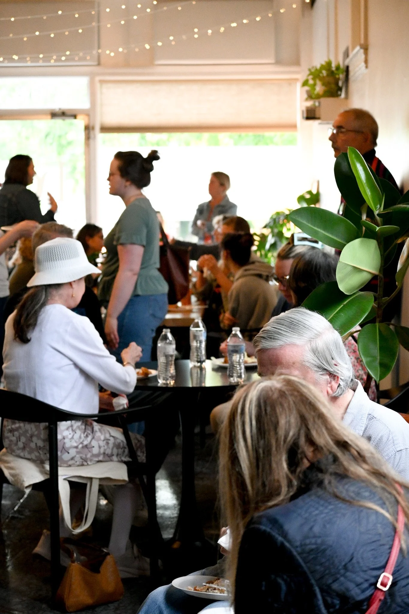 A group of people gathered in a well-lit indoor space, engaging in conversation and dining. The room is decorated with string lights and contains several potted plants. Some individuals are seated at tables with water bottles and plates of food, while others are standing and chatting.