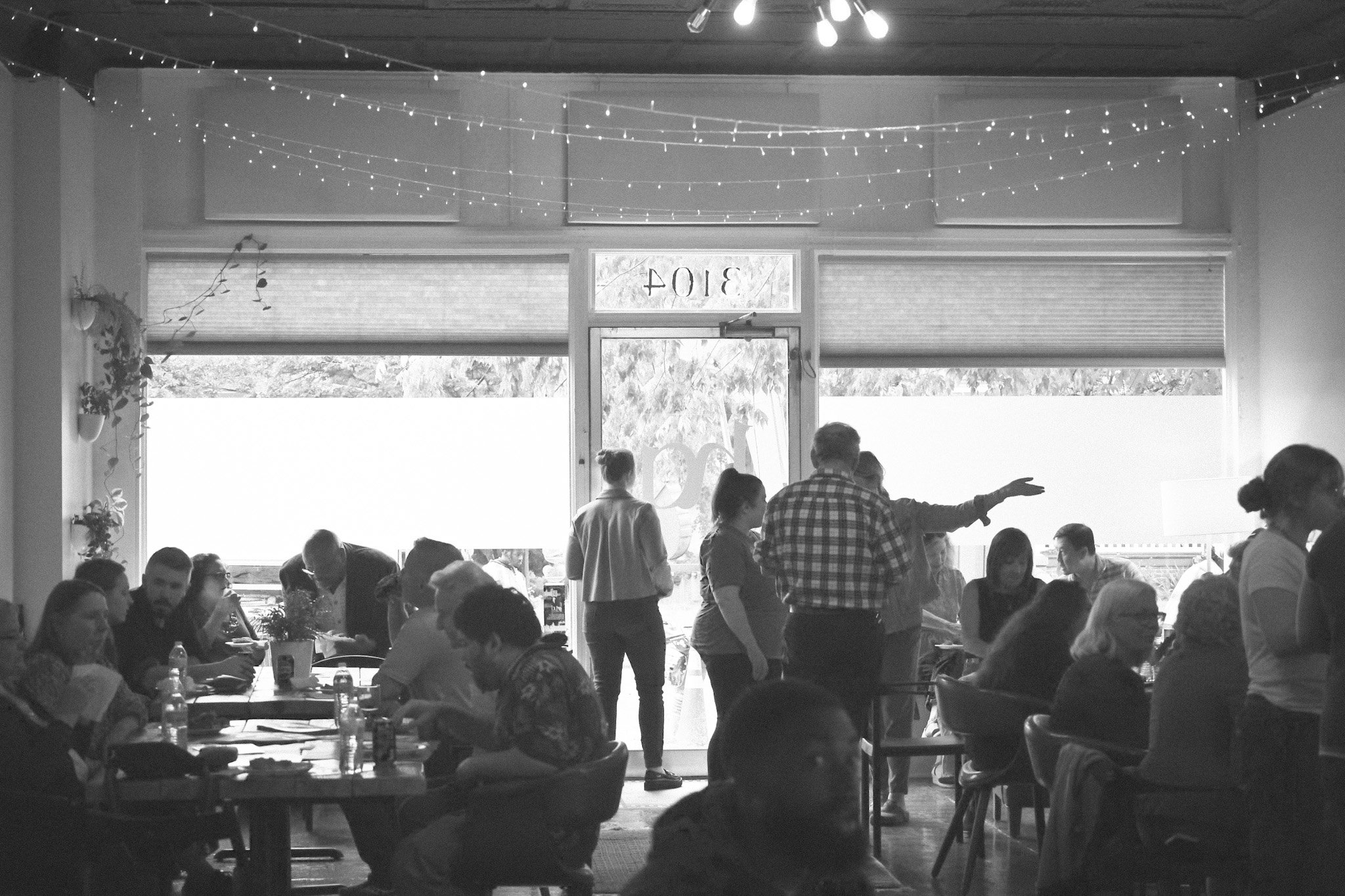 A group of people sitting at tables inside a cafe or restaurant, with some standing and talking. The room has large windows, string lights hanging from the ceiling, and a door in the background with graffiti on it. The scene is in black and white.