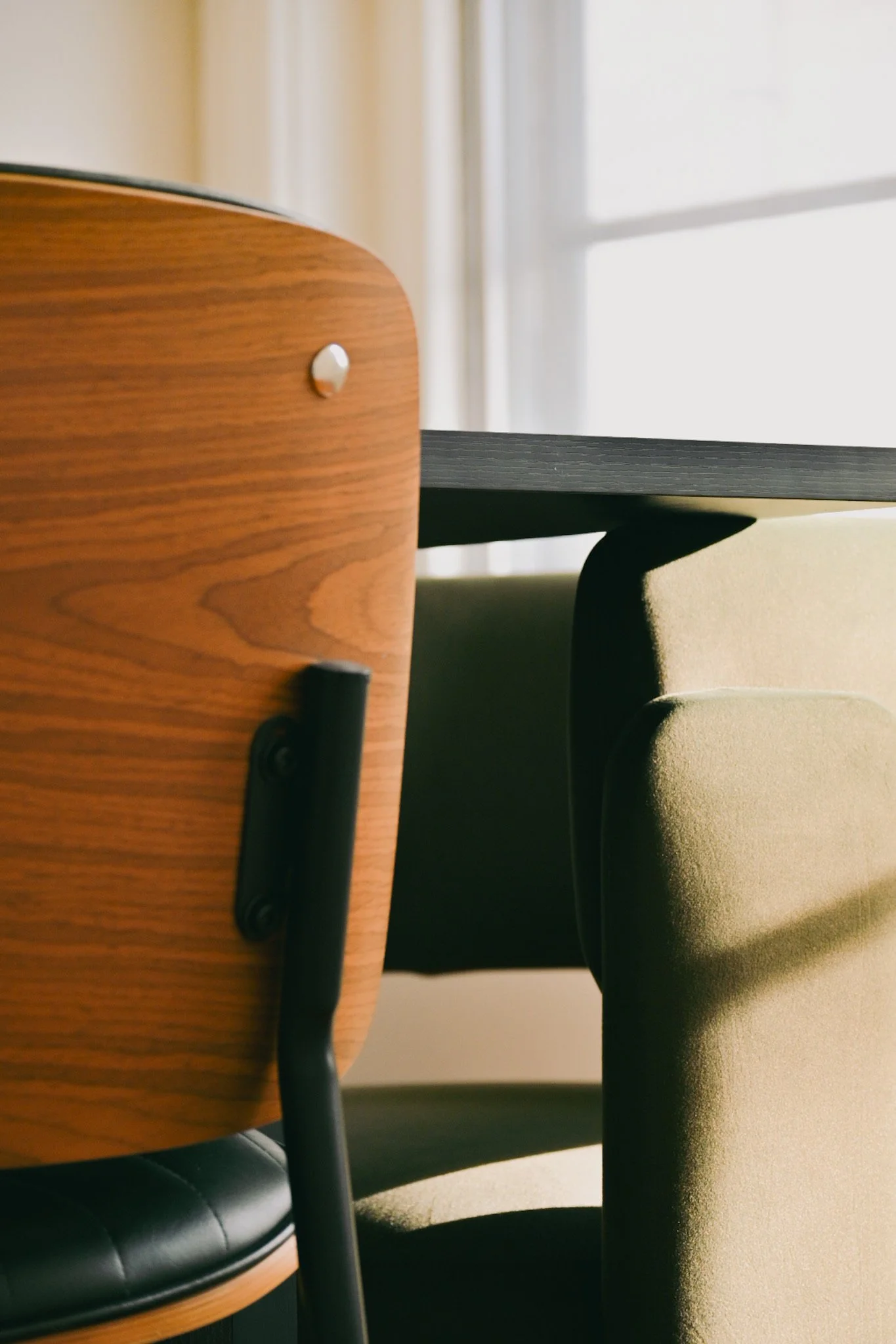 Close-up of a wooden chair with black metal legs, positioned next to a beige cushioned armchair near a window with white curtains, in a softly lit room.
