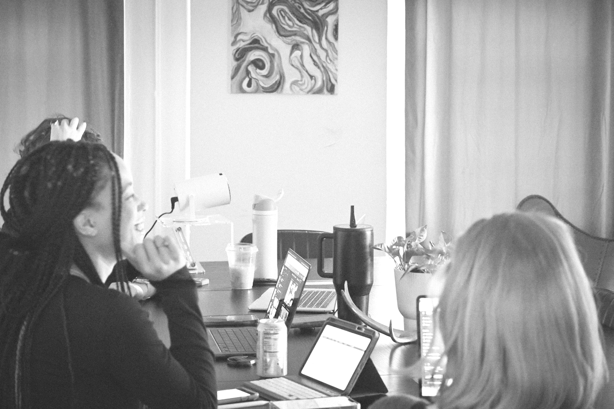 A group of four people working on laptops in an office or study space. One woman is in the foreground with glasses and braided hair, typing on a laptop. The others are partially visible, with one hand on her head. There are drinks and an electronic device on the table, with a wall and a decorative painting in the background.