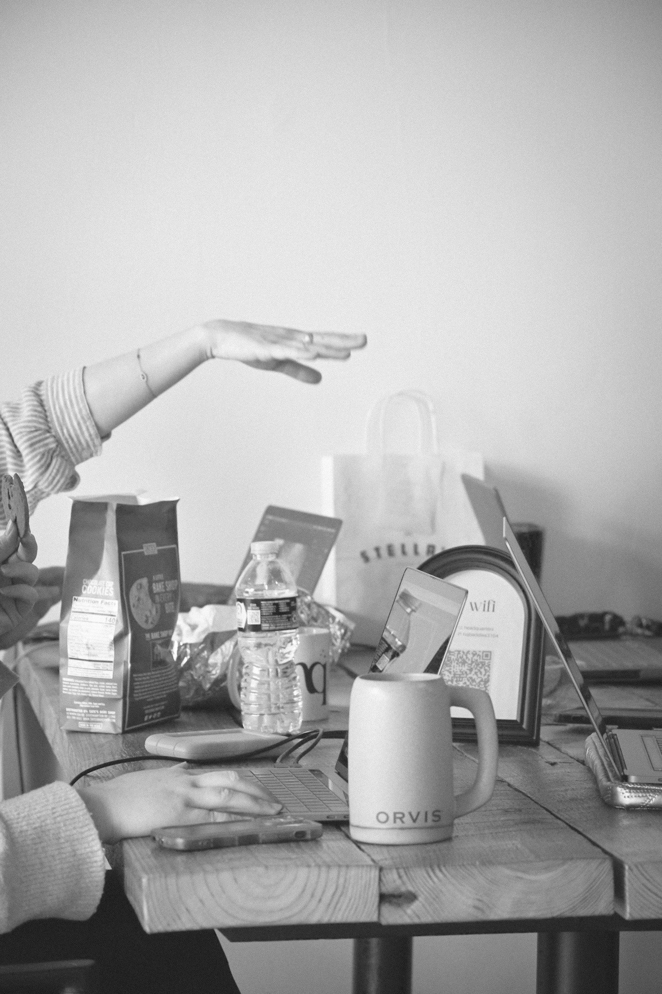A workspace with a person's hand typing on a laptop, surrounded by snacks, water bottles, a coffee mug labeled 'ORVIS', and various electronic devices on a wooden table.